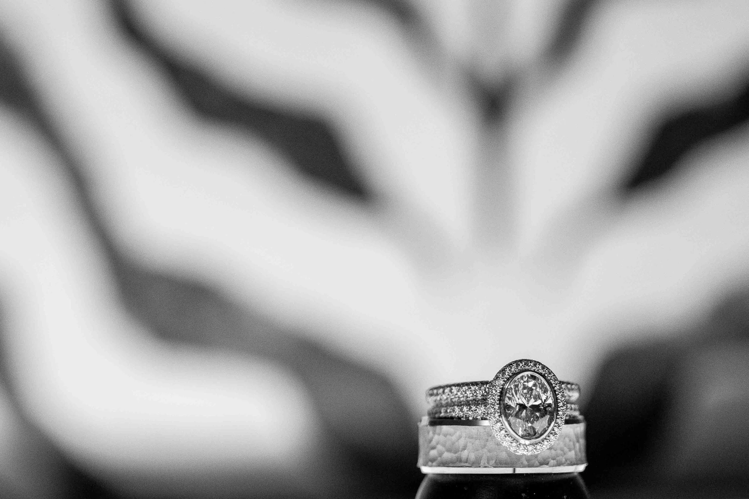 Close-up of a diamond engagement ring with a large central stone and a band adorned with smaller diamonds, placed on a ring holder with a blurred hand holding a small object in the background.