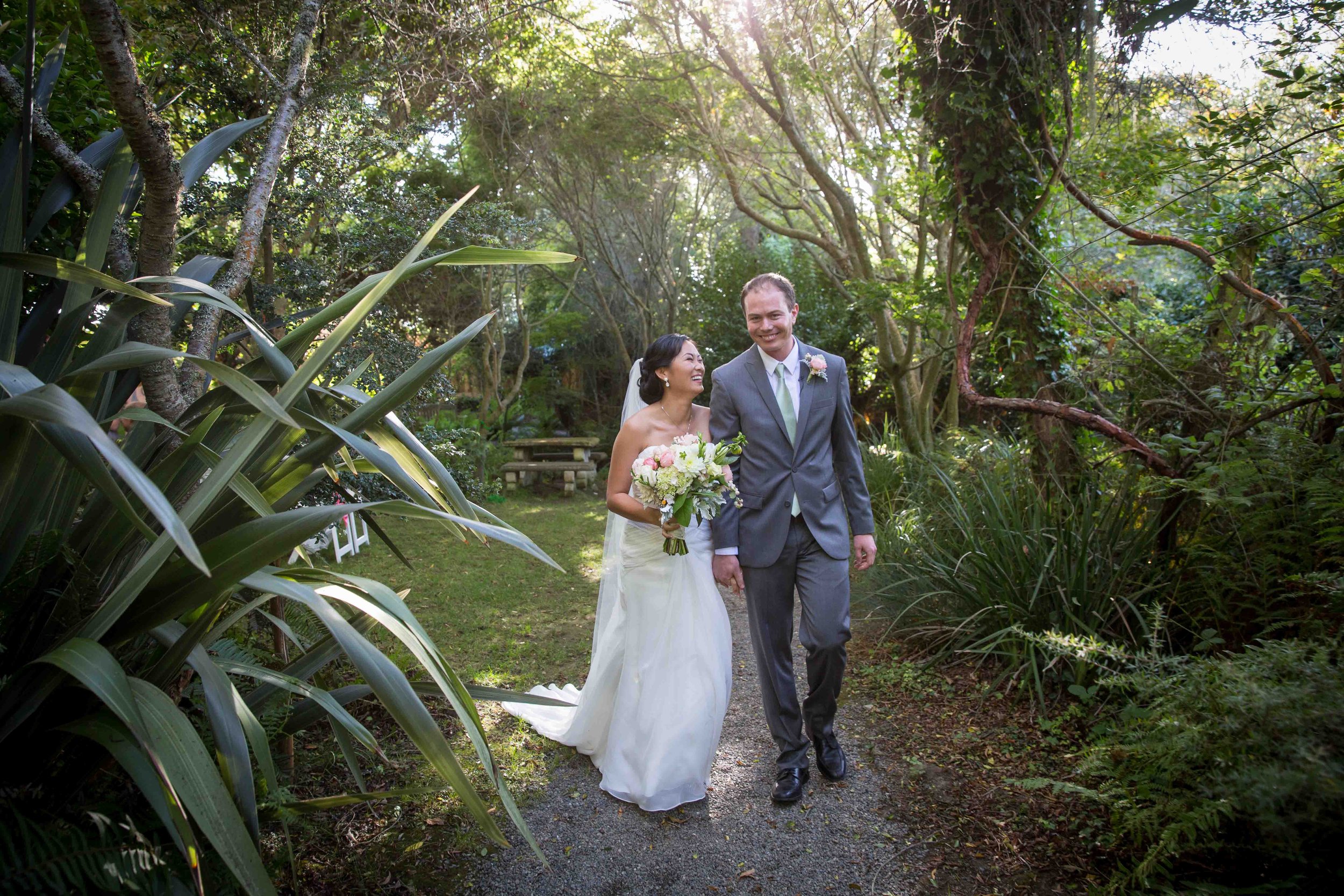 A bride and groom walking hand in hand along a garden path, smiling at each other. The bride is holding a bouquet of flowers, wearing a white wedding gown and veil. The groom is dressed in a gray suit with a light green tie. The garden is lush and gr