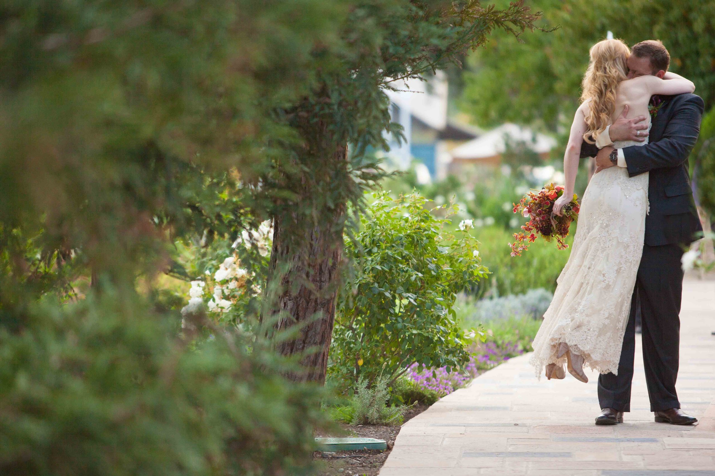 A bride and groom hugging outdoors on a wedding day, with greenery and flowers around them.