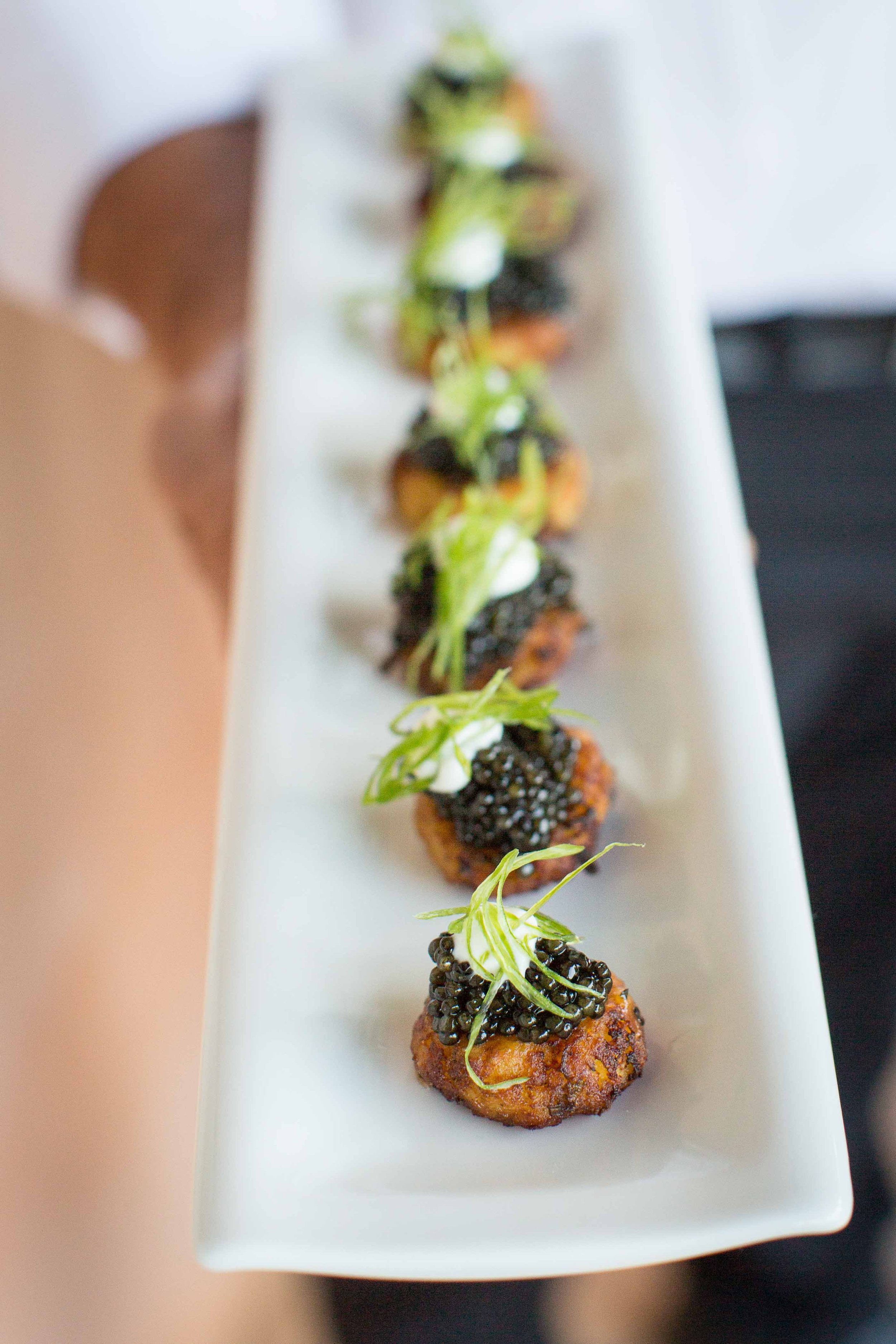 Close-up of a white rectangular plate with five small appetizers topped with caviar, garnished with green herbs and dollops of mayonnaise, arranged in a row.