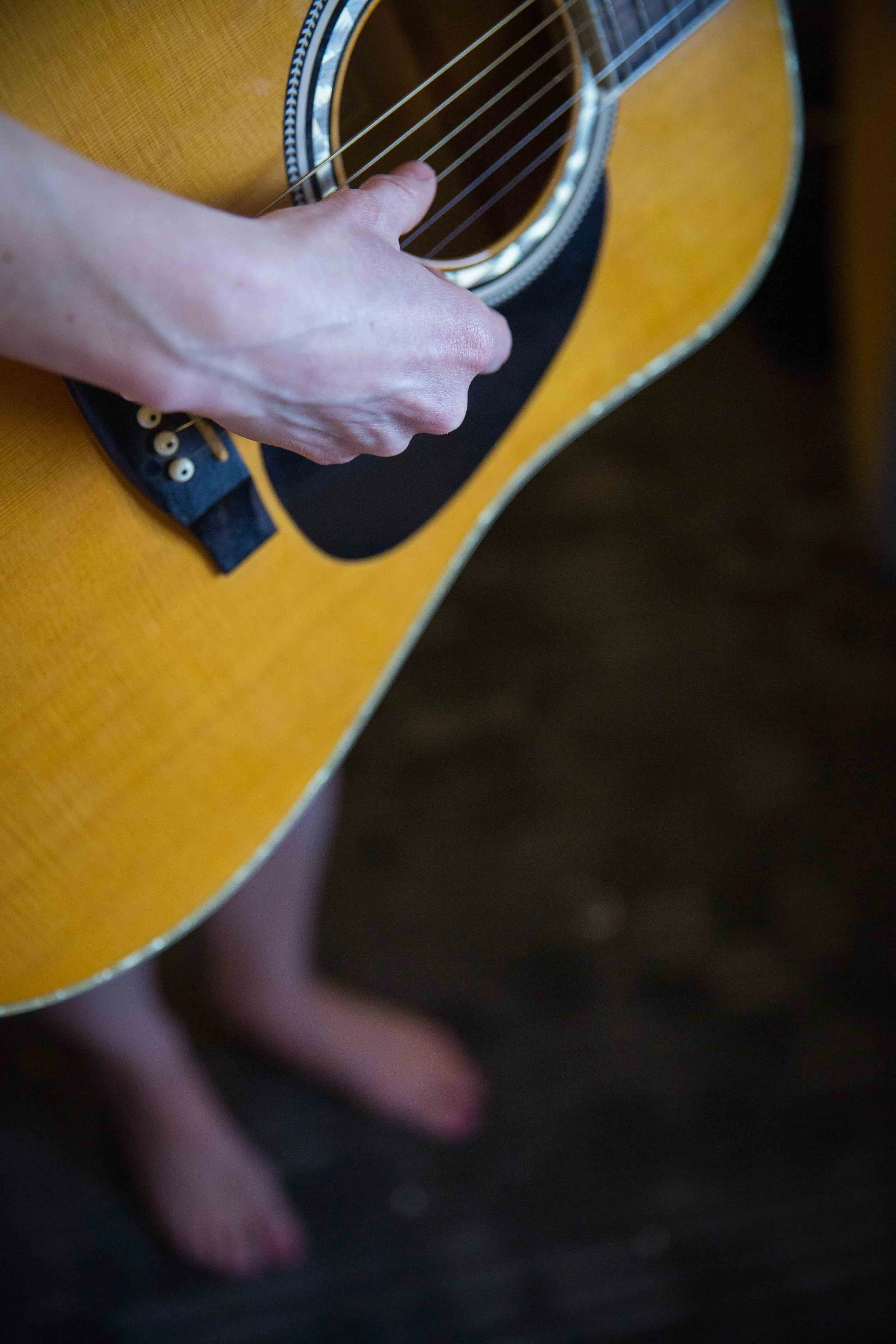 Close-up of a person’s hand strumming an acoustic guitar, with their feet visible in the background.