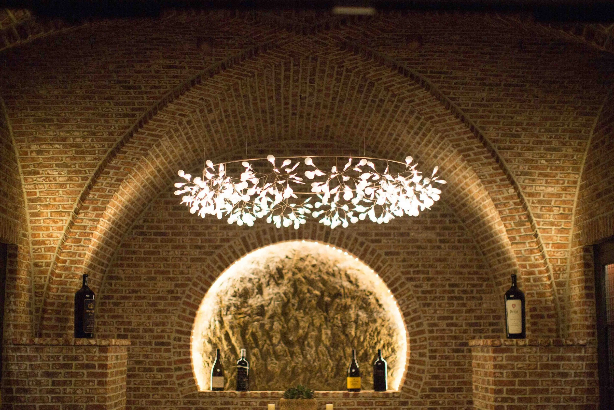 Interior of a brick-walled wine cellar with an arched ceiling, illuminated by a decorative chandelier resembling branches with leaves, and bottles of wine on a ledge.