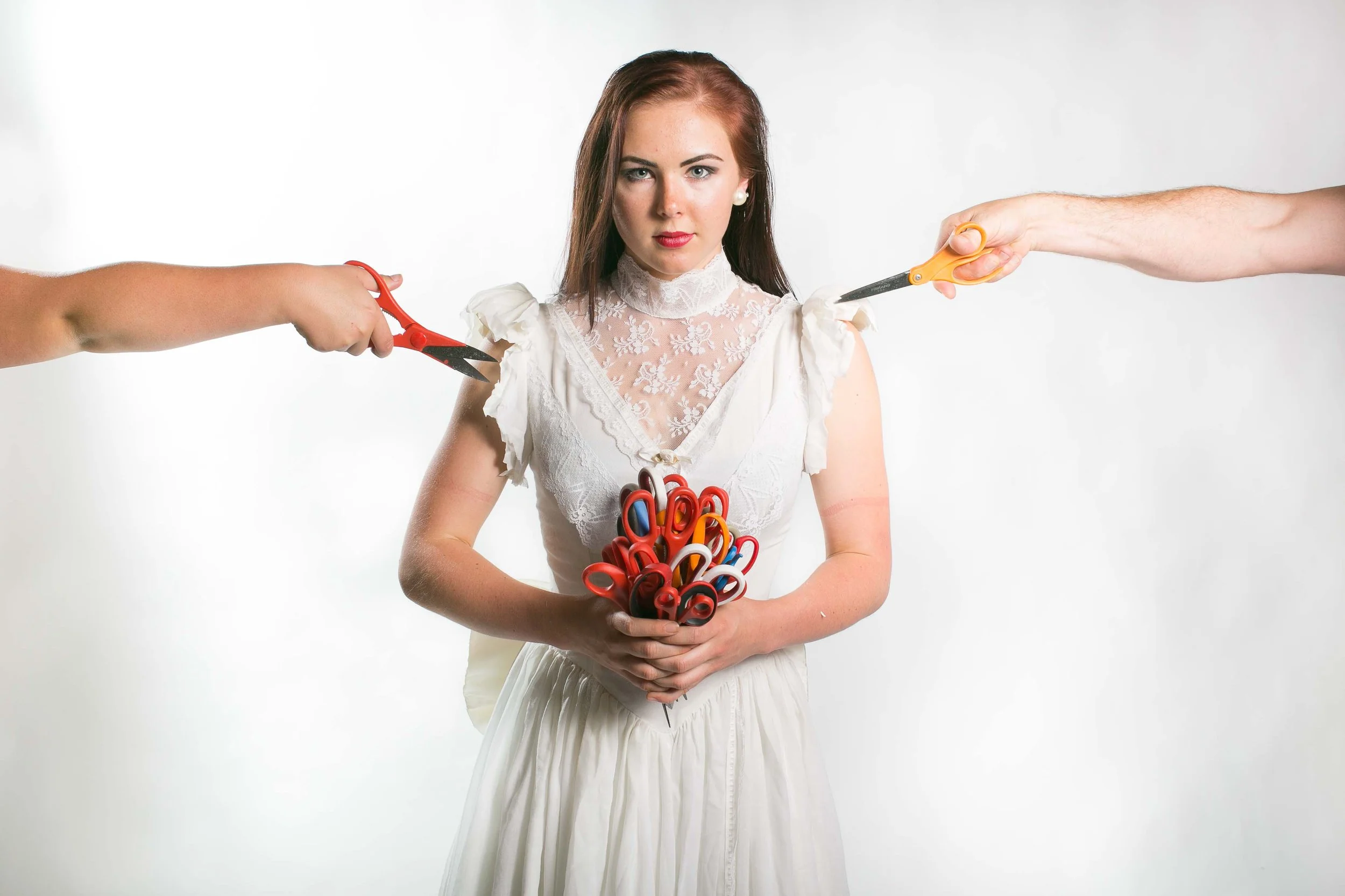 A woman in a white dress holds a bunch of colorful scissors. Two other people are extending their arms with scissors toward her, creating an appearance of a tense standoff.