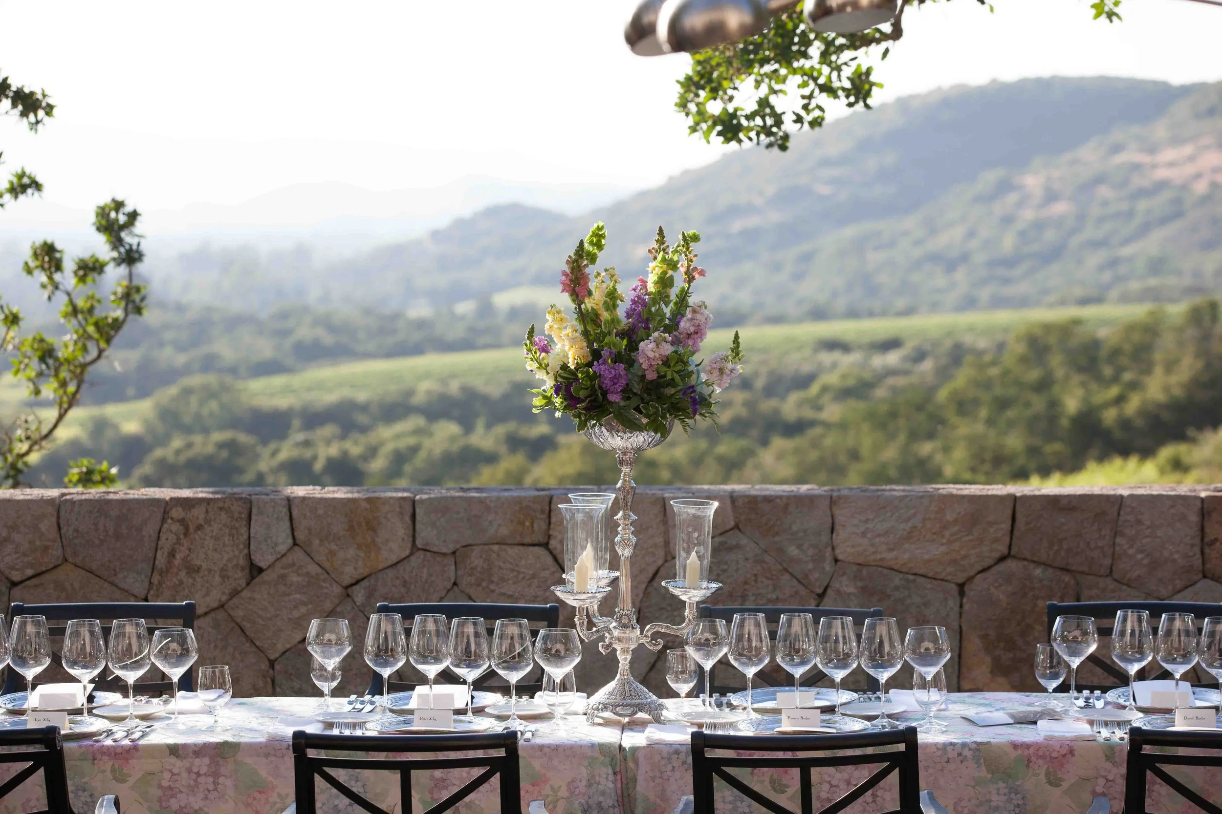 Elegant outdoor dining table set with glasses and a floral centerpiece, overlooking a scenic mountain landscape.