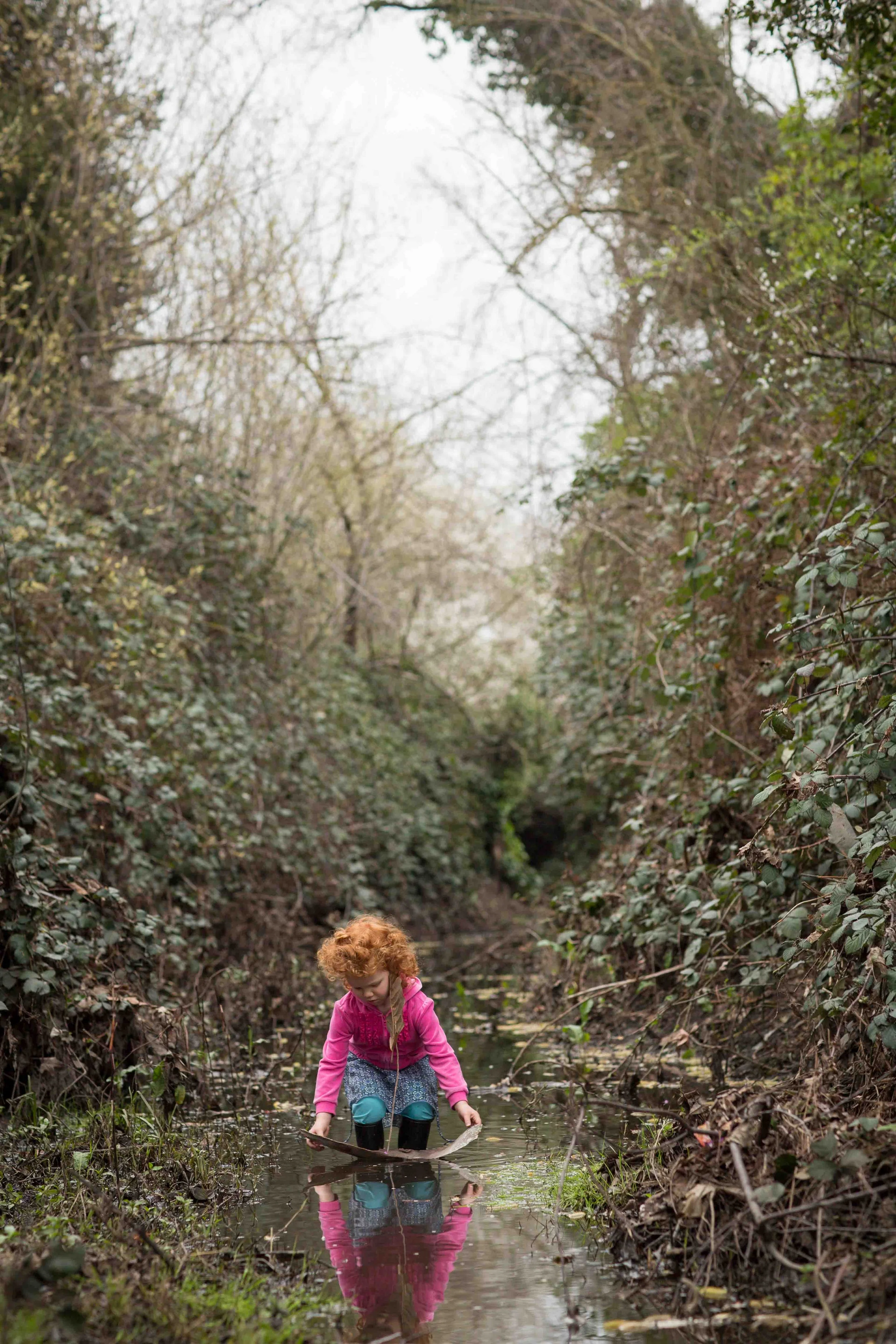 A young girl with curly red hair in a pink jacket and blue pants is playing in a shallow muddy stream in a wooded area.