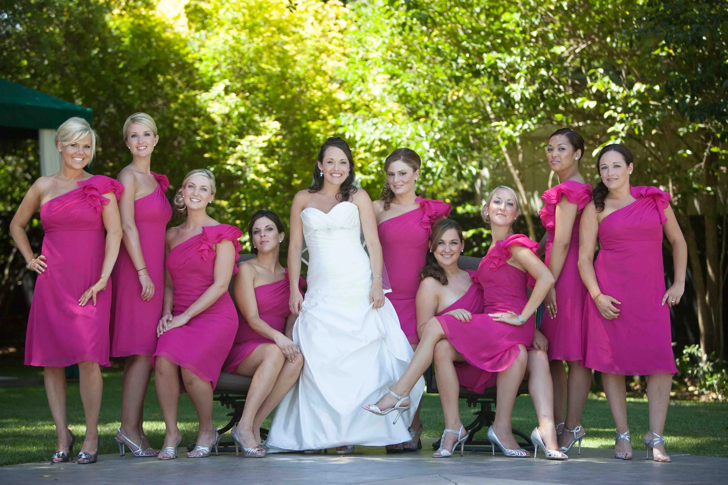 Group of women, including a bride in a white wedding dress, with bridesmaids in bright pink dresses, posing outdoors in a garden.