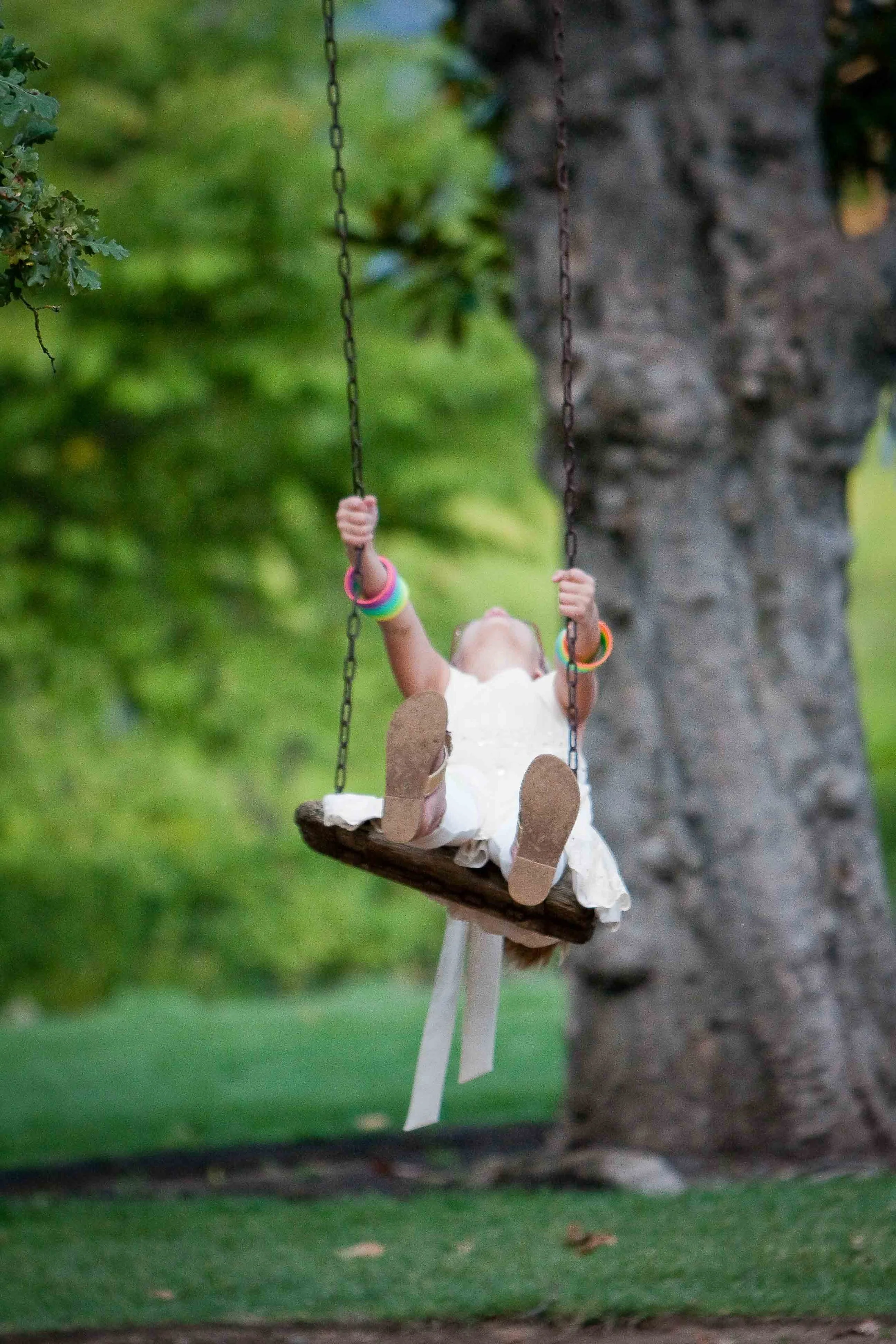 A young girl wearing white clothes and colorful bracelets is swinging on a wooden swing hanging from a tree in a park.