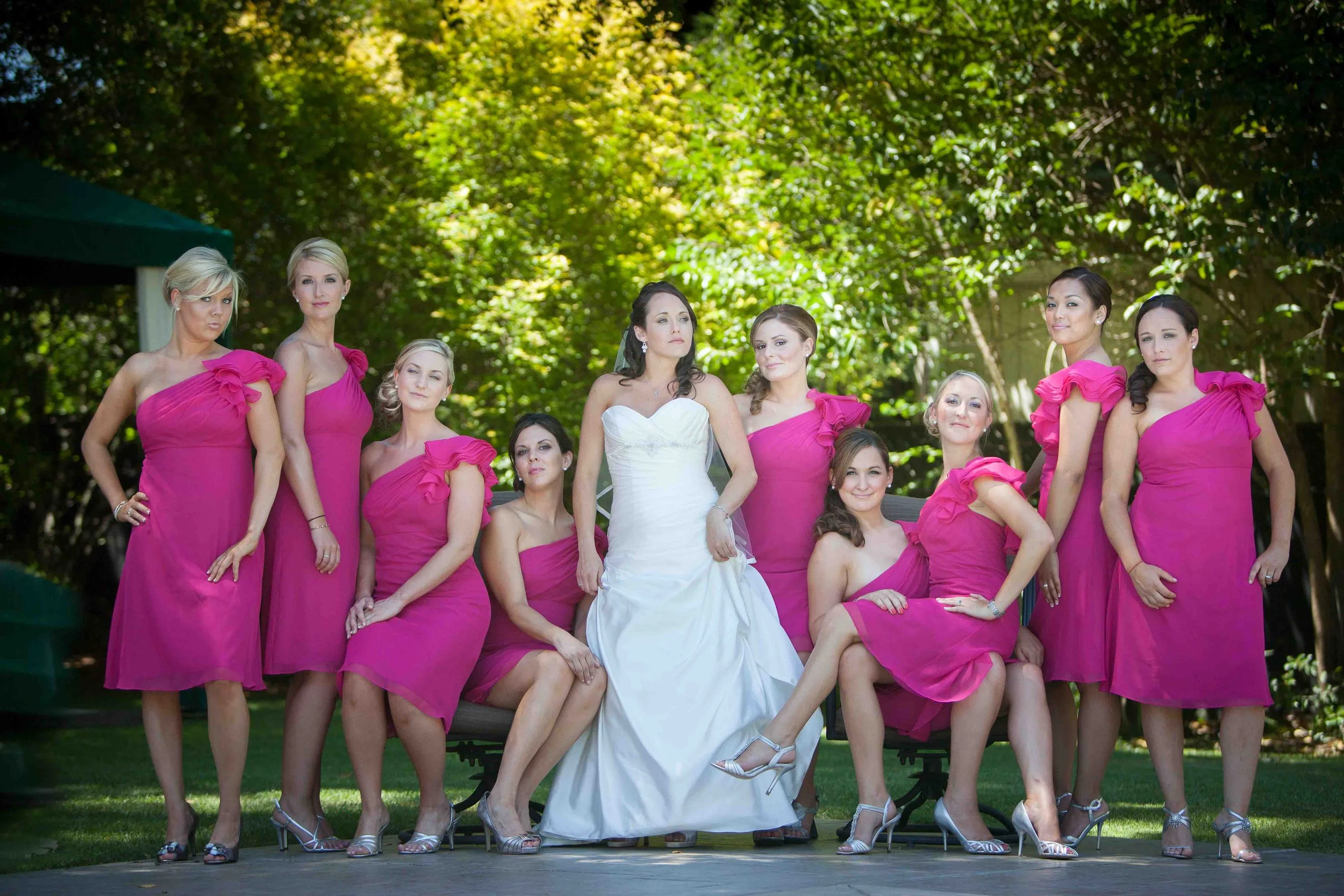 A bride in a white wedding dress sitting on a bench surrounded by nine bridesmaids in pink dresses, outdoors with green trees in the background.
