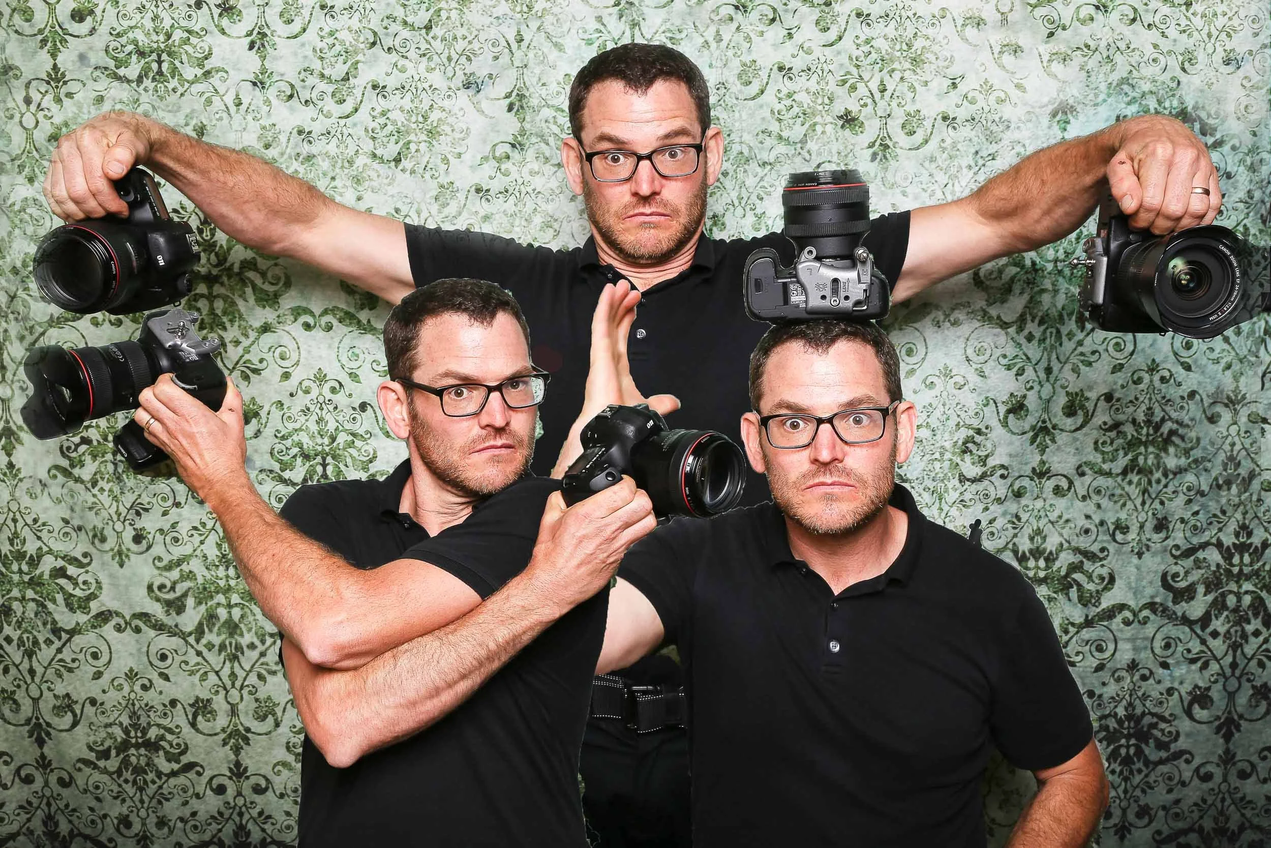 A man with glasses and a black shirt posing with four different cameras in front of a decorative green patterned background.