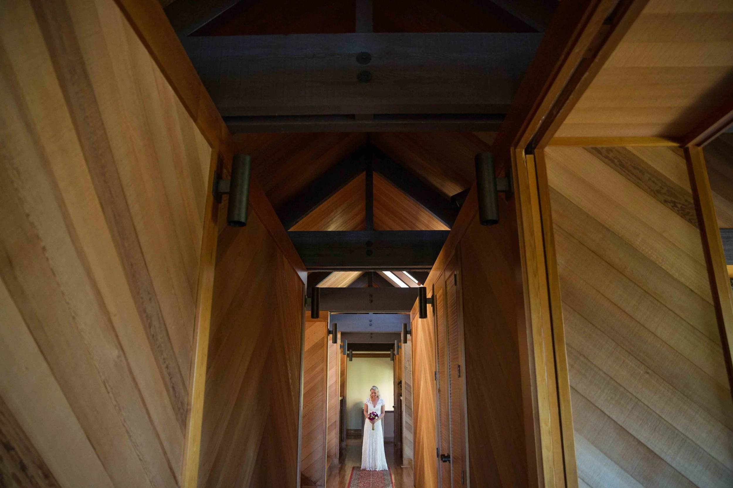 A woman in a white wedding dress holding a bouquet standing at the end of a narrow, wood-paneled hallway with a triangular-patterned ceiling.