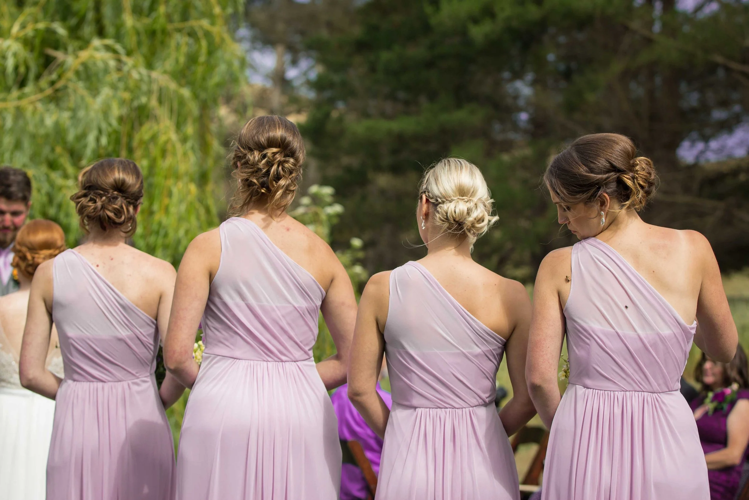 Four women in matching lavender bridesmaid dresses standing outdoors with trees in the background, seen from behind.