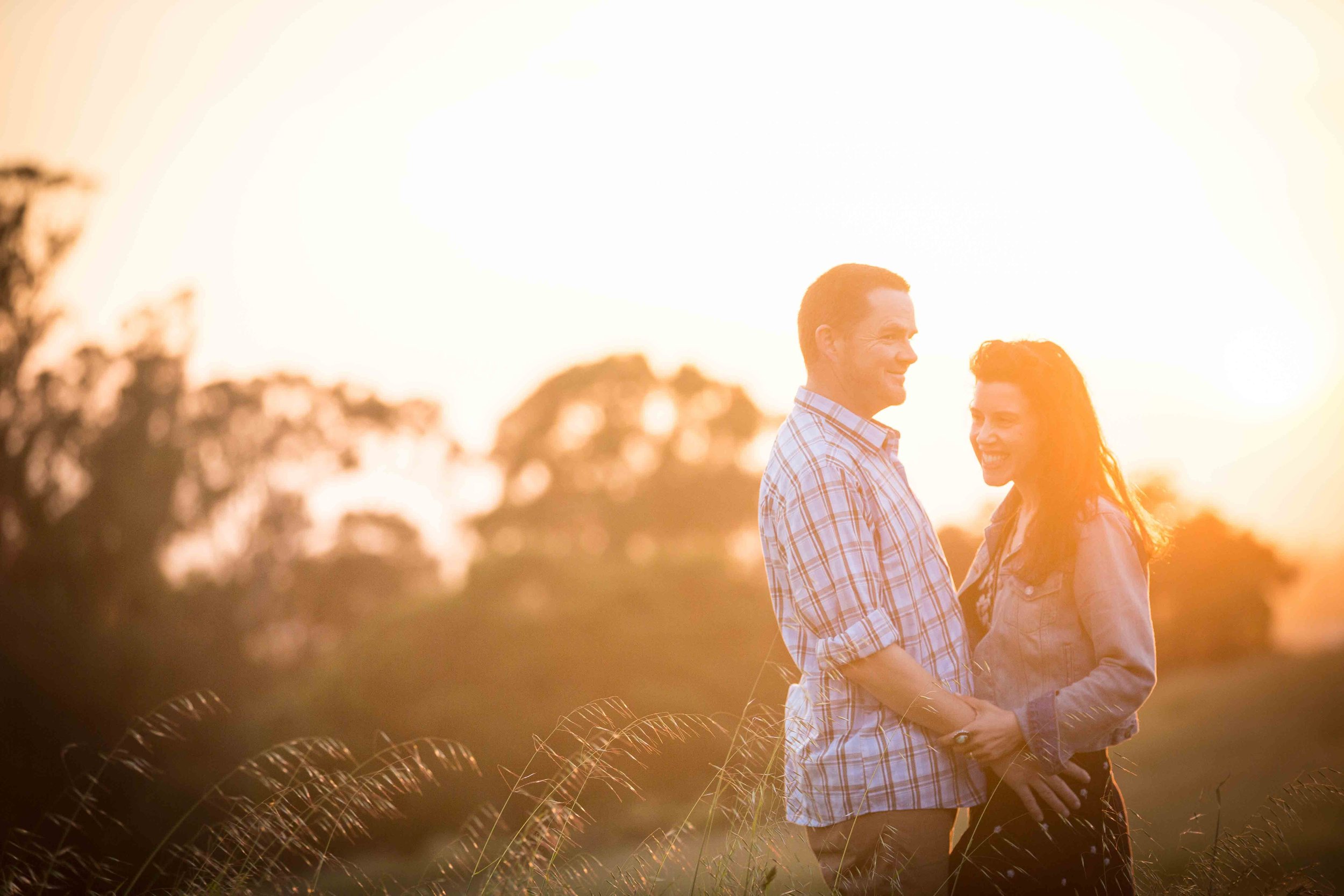 A couple standing outdoors in a field during sunset, smiling and holding hands.