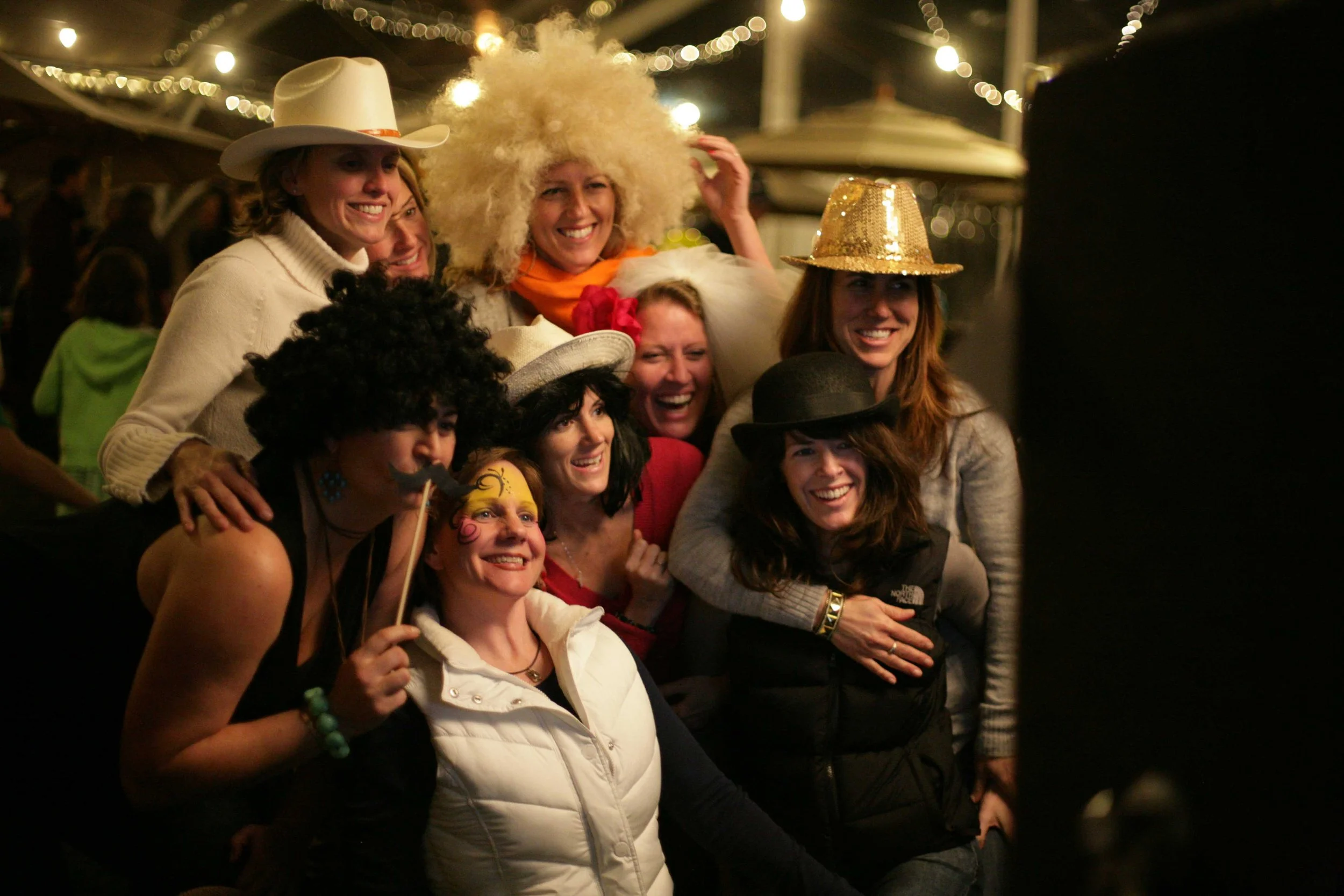 Group of women in costumes and hats, smiling and posing together at a night event under string lights.
