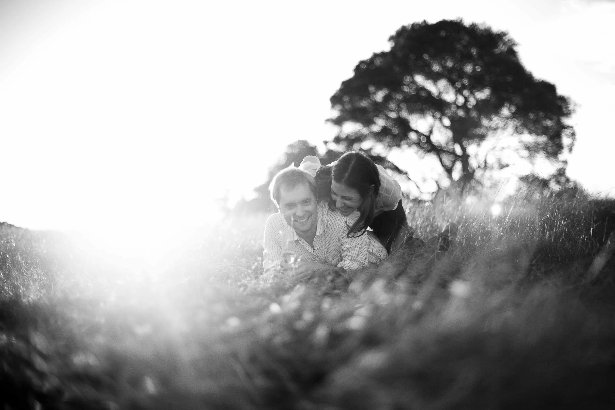 A black and white photo of a smiling couple lying on the grass outdoors, with a large tree in the background. The woman is leaning over the man, and they appear happy and playful.