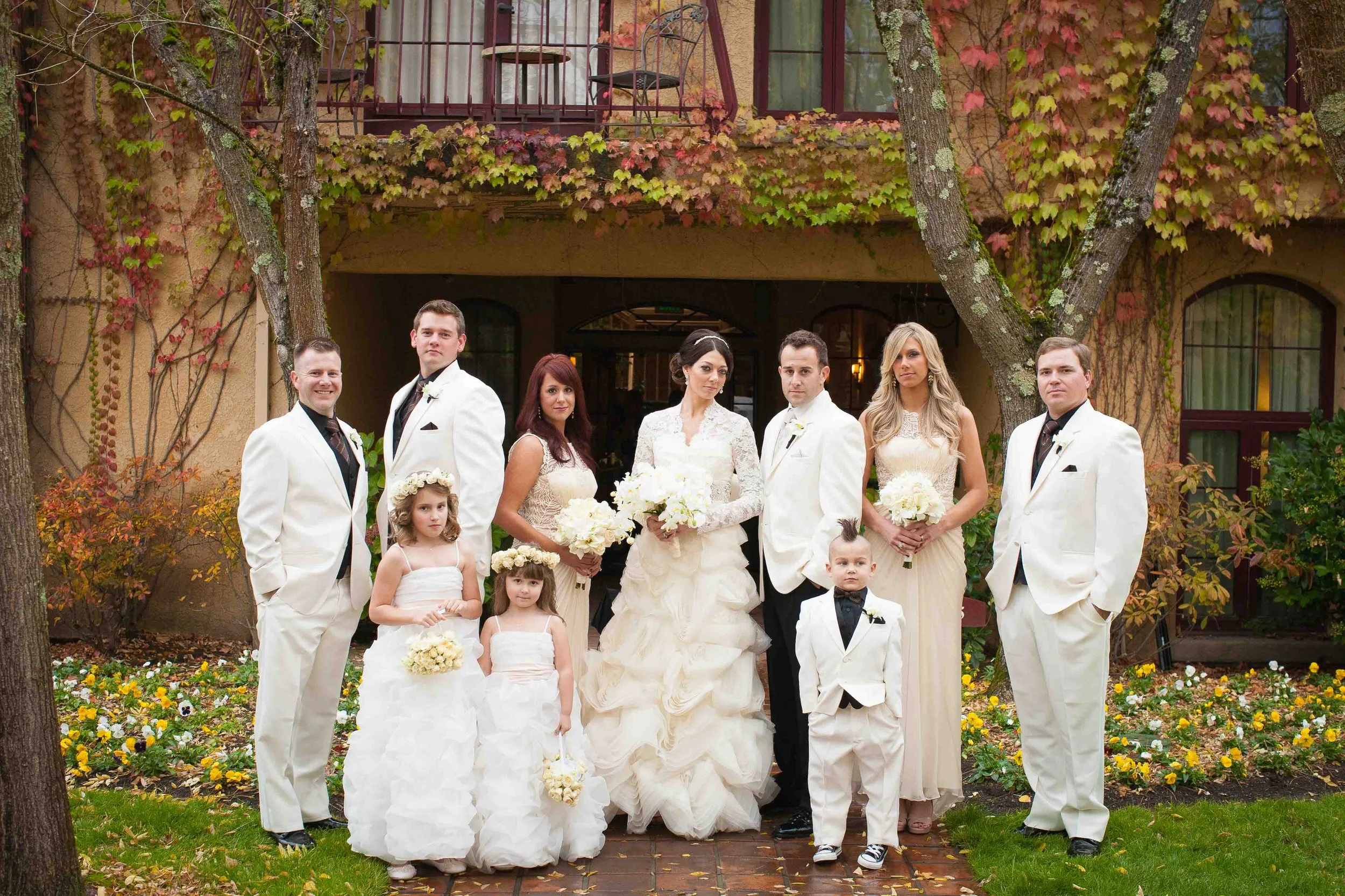 A wedding party outside in a garden with autumn leaves on trees and flowers on the ground. The bride and groom are in the center, surrounded by bridesmaids and groomsmen, all dressed in formal attire, with children in white dresses and tuxedos.