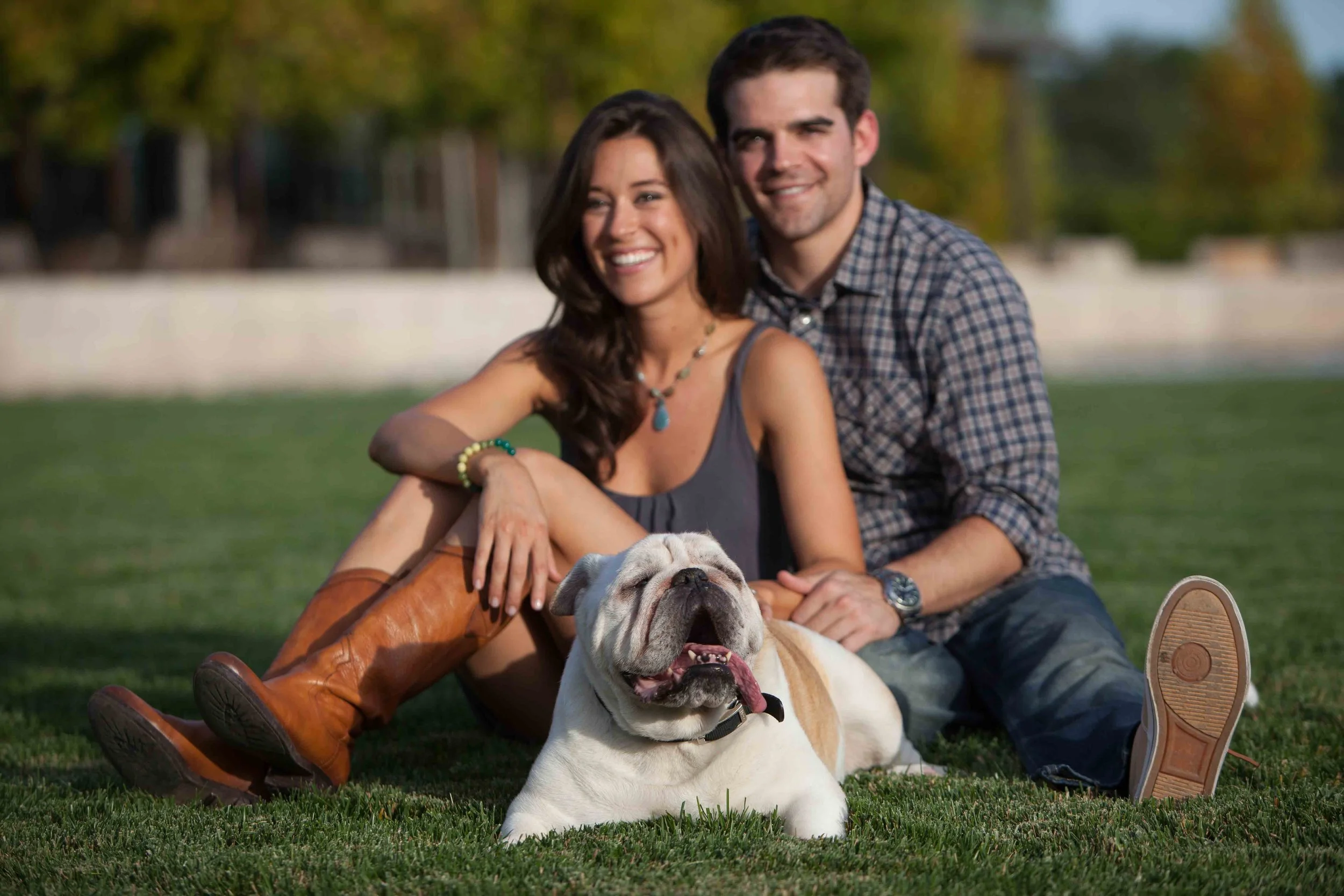 A happy couple sitting on the grass with their bulldog in a park, smiling at the camera.