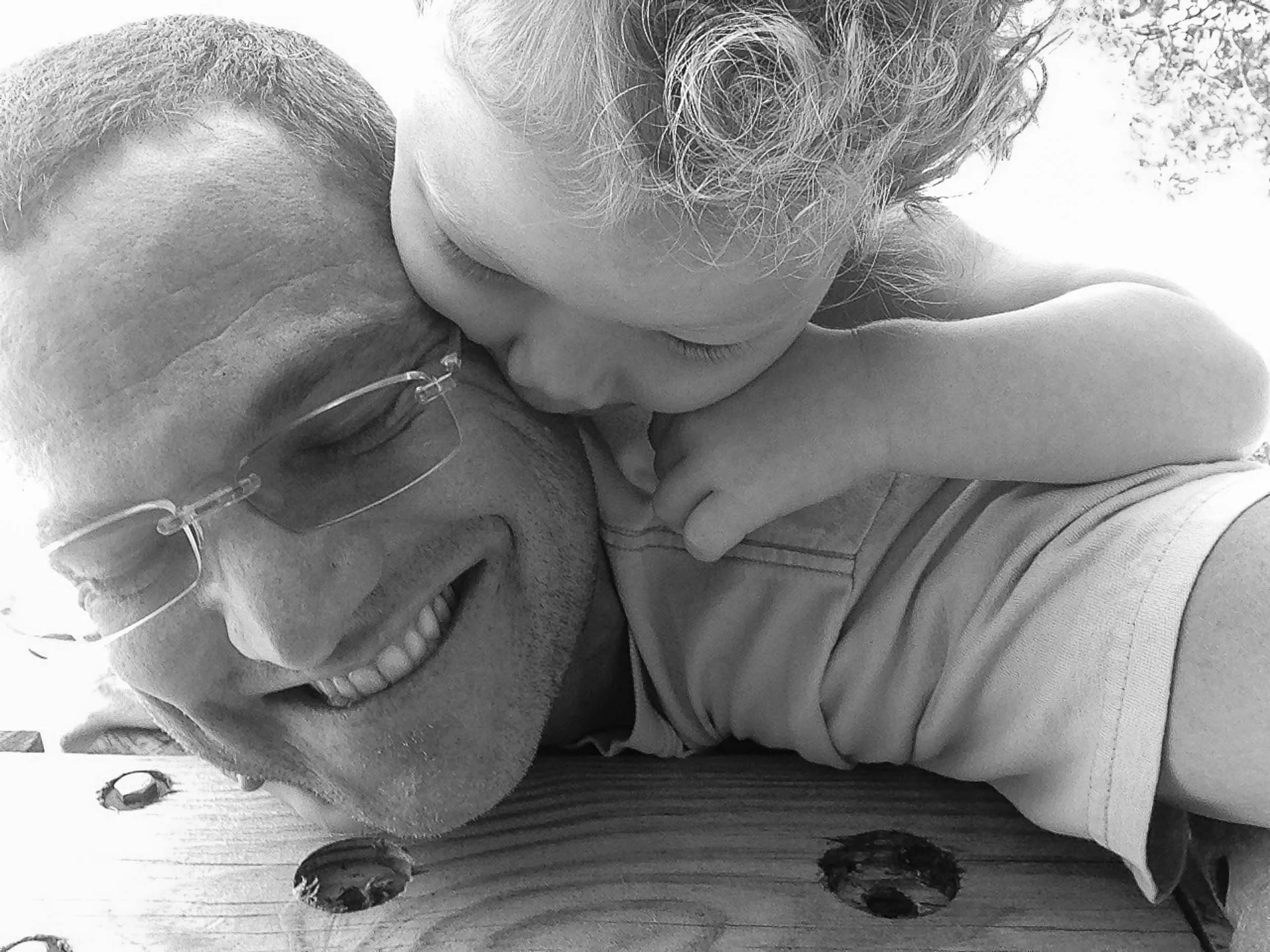 A man smiling with glasses and a child with curly hair kissing his cheek, both lying on a wooden surface.