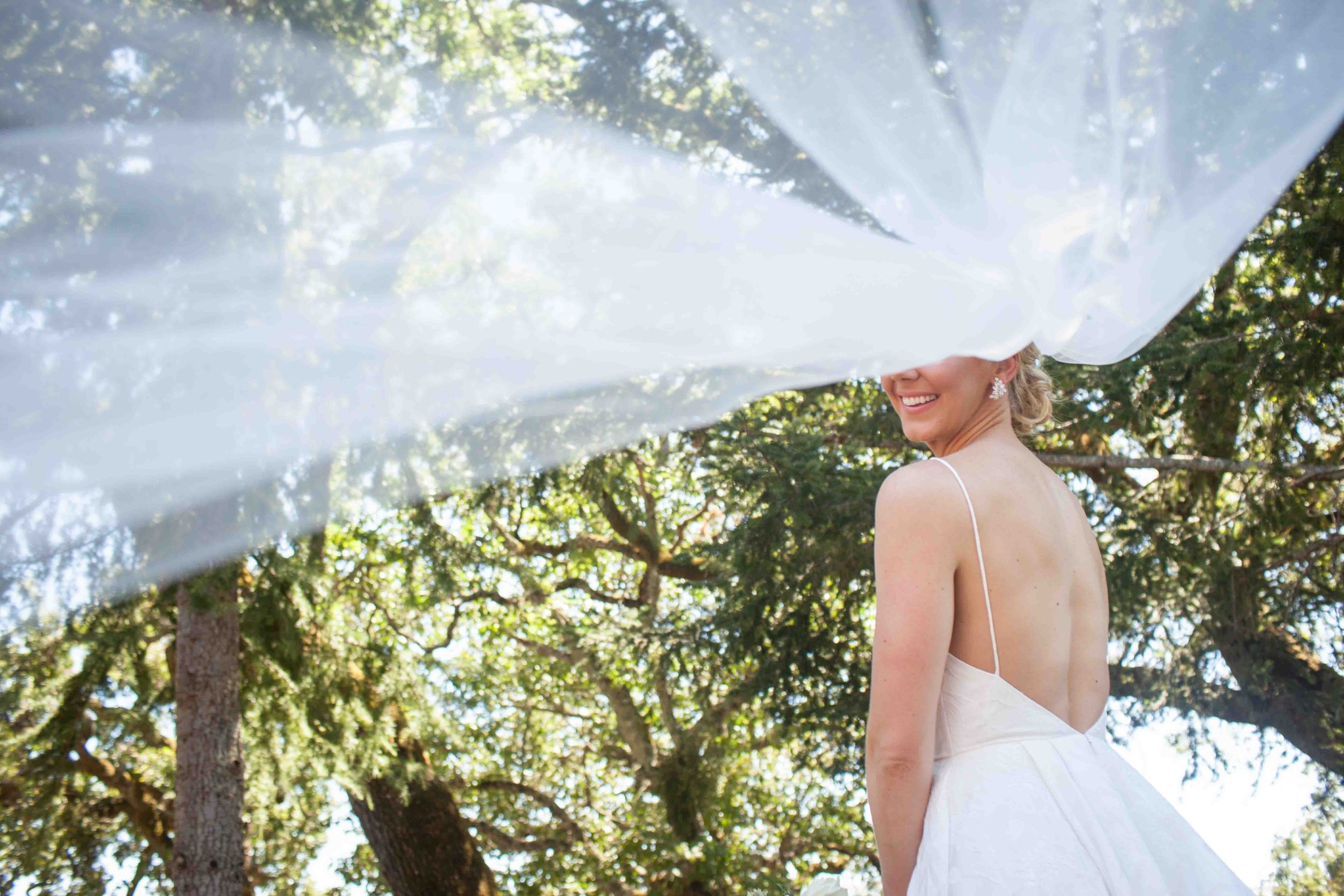 A bride in a white wedding dress and jewelry smiling, with trees and sunlight in the background, partly obscured by a large sheer white fabric.