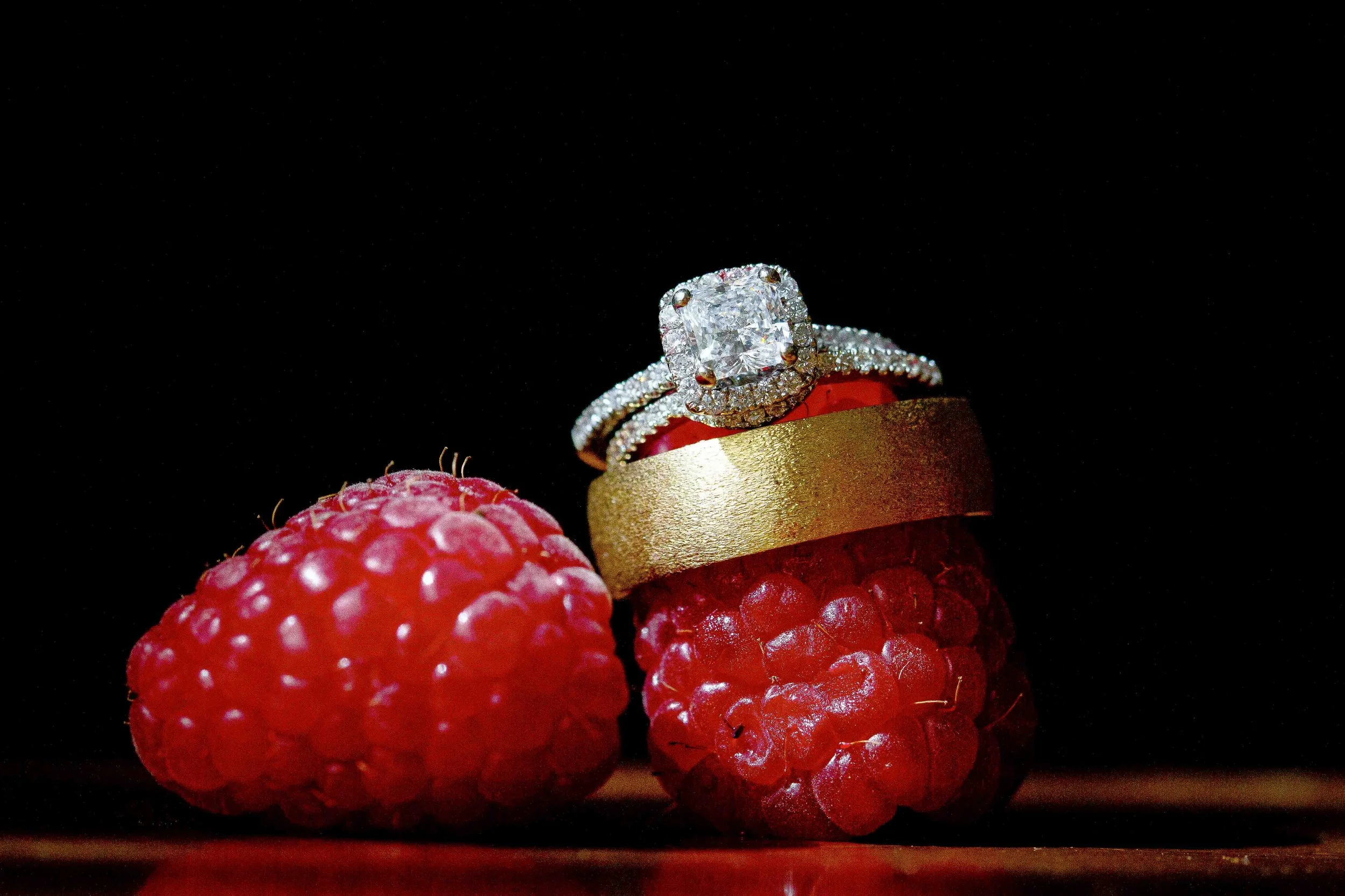 Close-up of two fresh raspberries and a gold ring with a large diamond on top, set against a dark background.