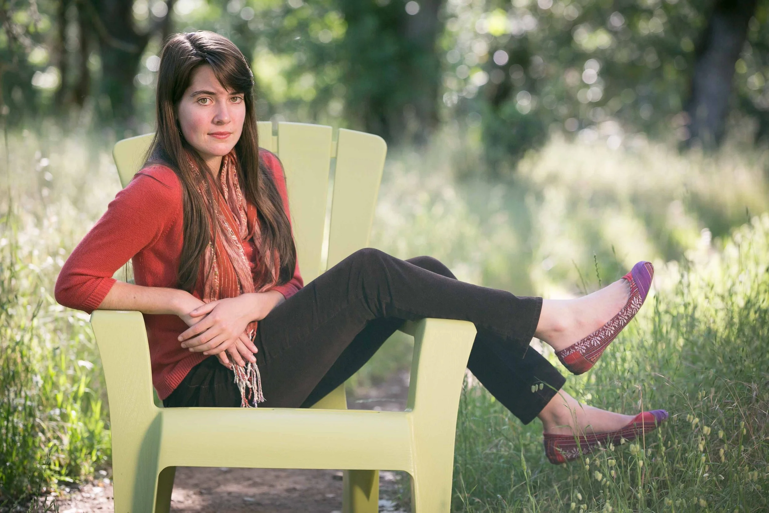 A woman sitting in a light green outdoor chair with her leg resting on the grass in a park, wearing a red top, black pants, and patterned red shoes. She has long dark hair and is looking at the camera.
