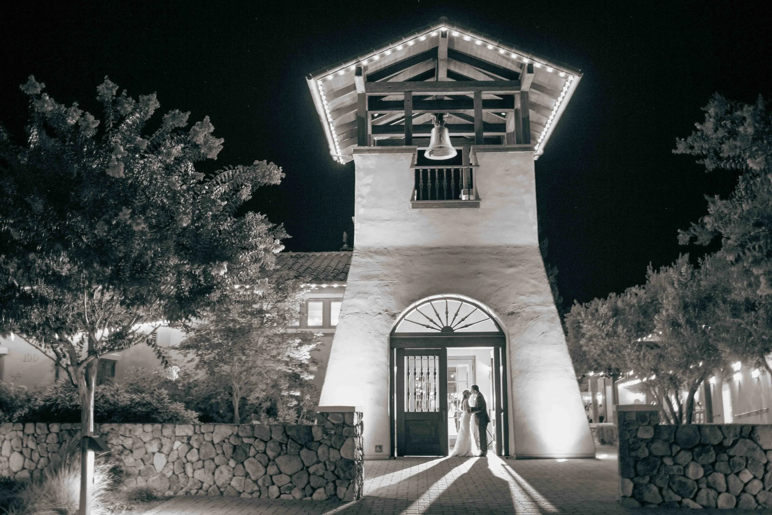 A couple getting married at night under a lit tower with trees and a stone wall nearby.