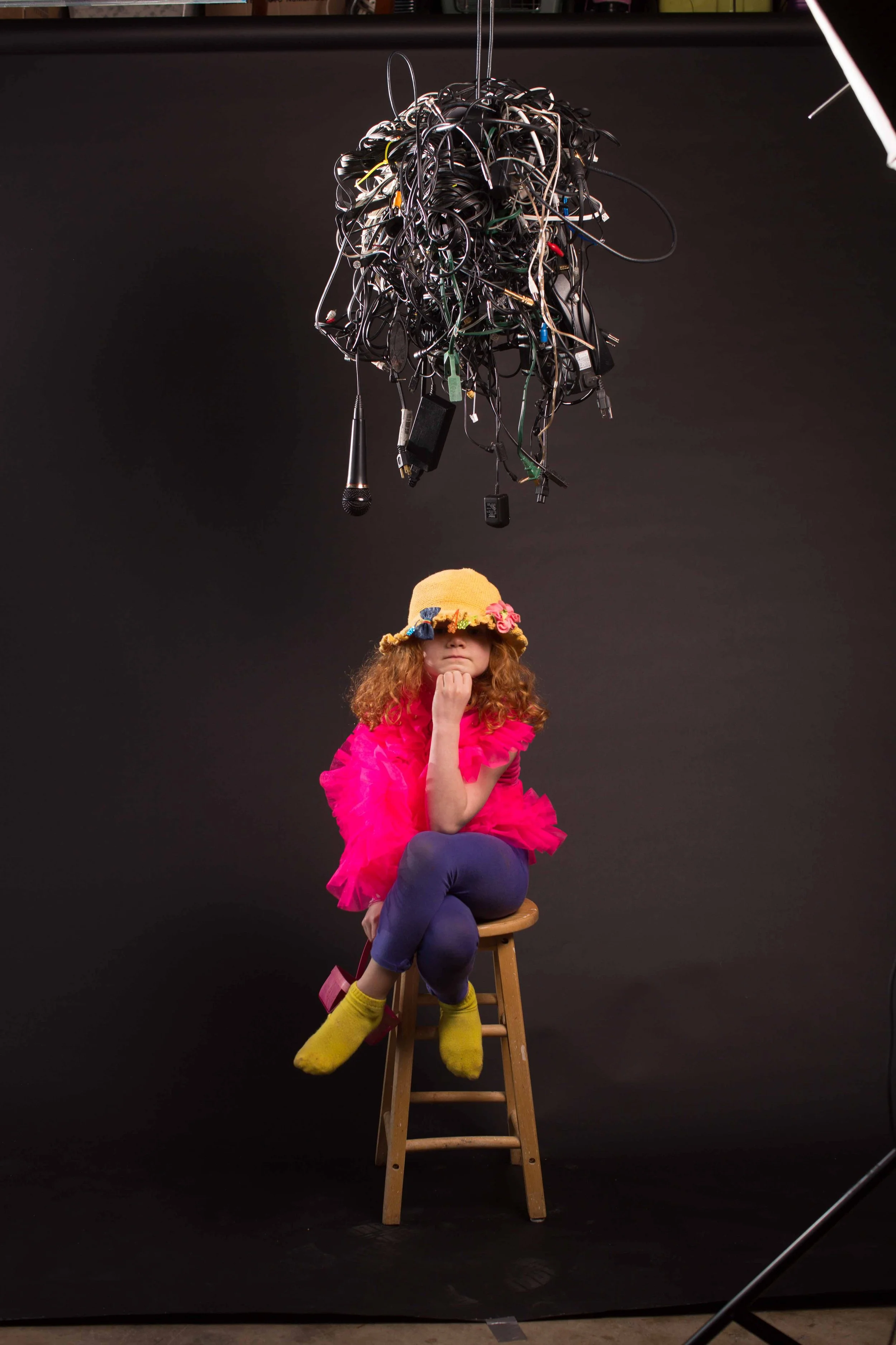 A young girl with curly red hair, wearing a yellow hat with a flower decoration, bright pink ruffled top, blue pants, and yellow socks, sits on a wooden stool with her chin resting on her hand, under a large tangle of black electrical wires hanging f