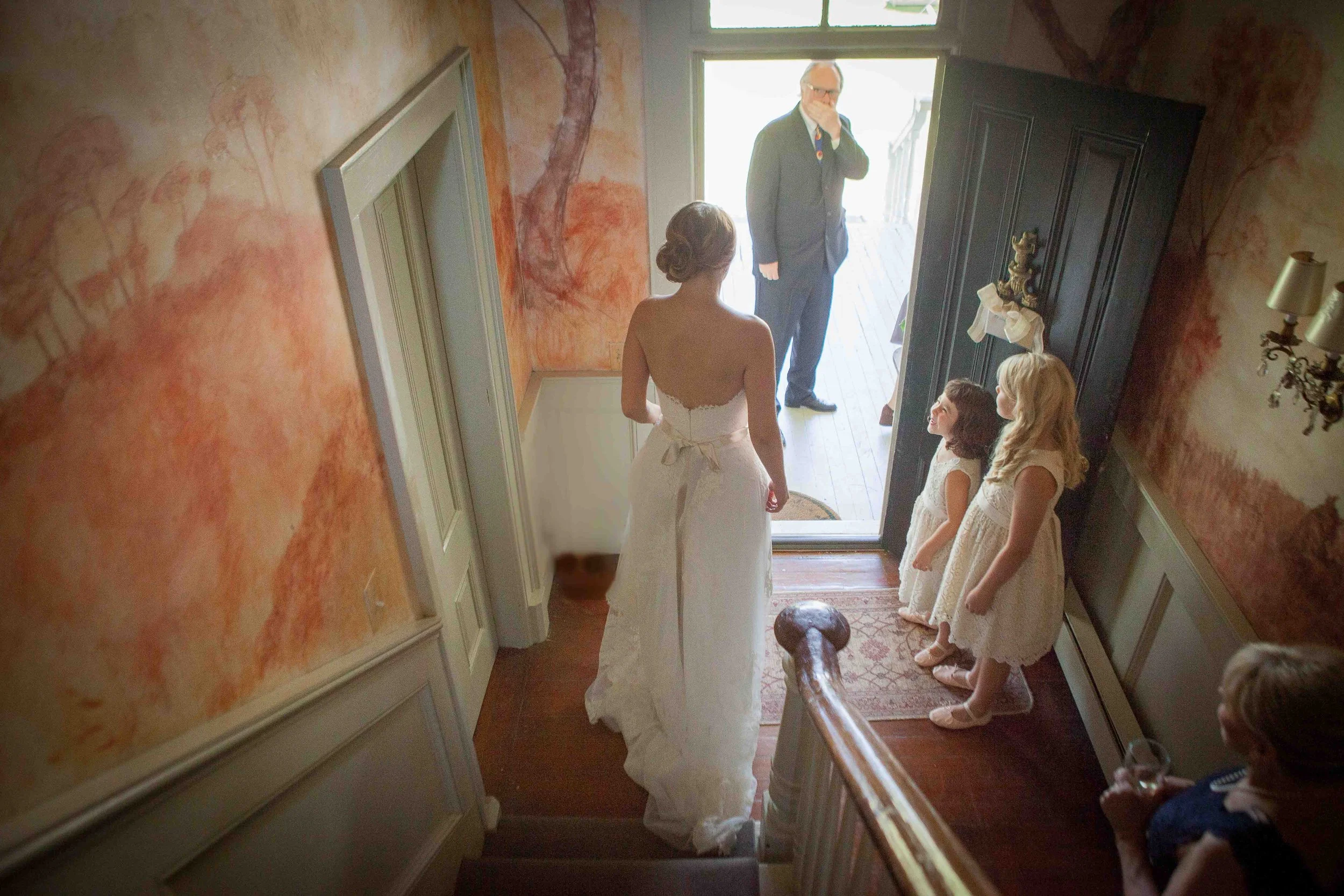 A bride in a strapless wedding gown standing at the top of a staircase facing an open door, with an older man and several flower girls in white dresses nearby. A woman seated at the bottom of the stairs is holding a glass, observing the scene.