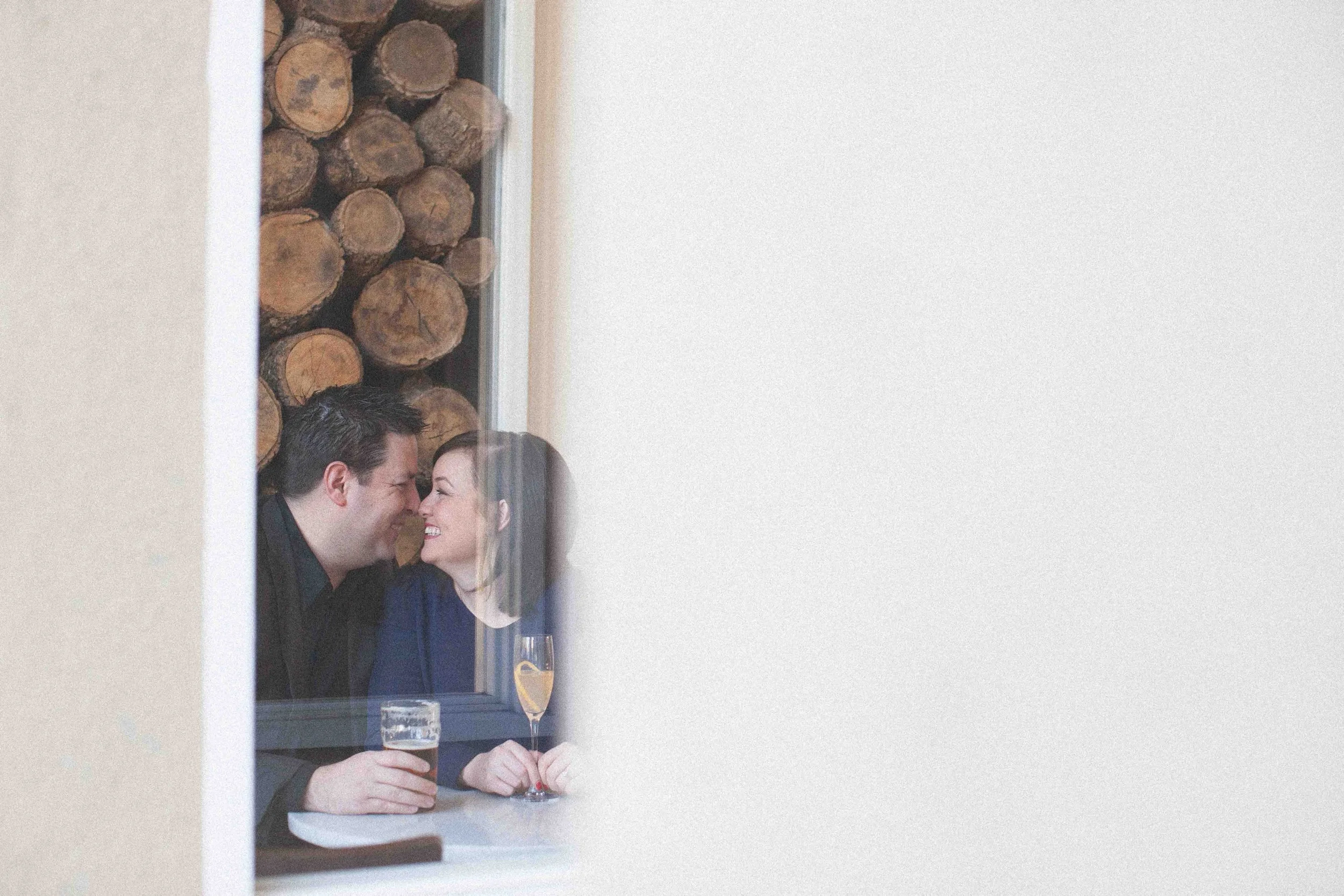 A couple making a face through a window, touching noses, with drinks on the table, and a stack of logs in the background.