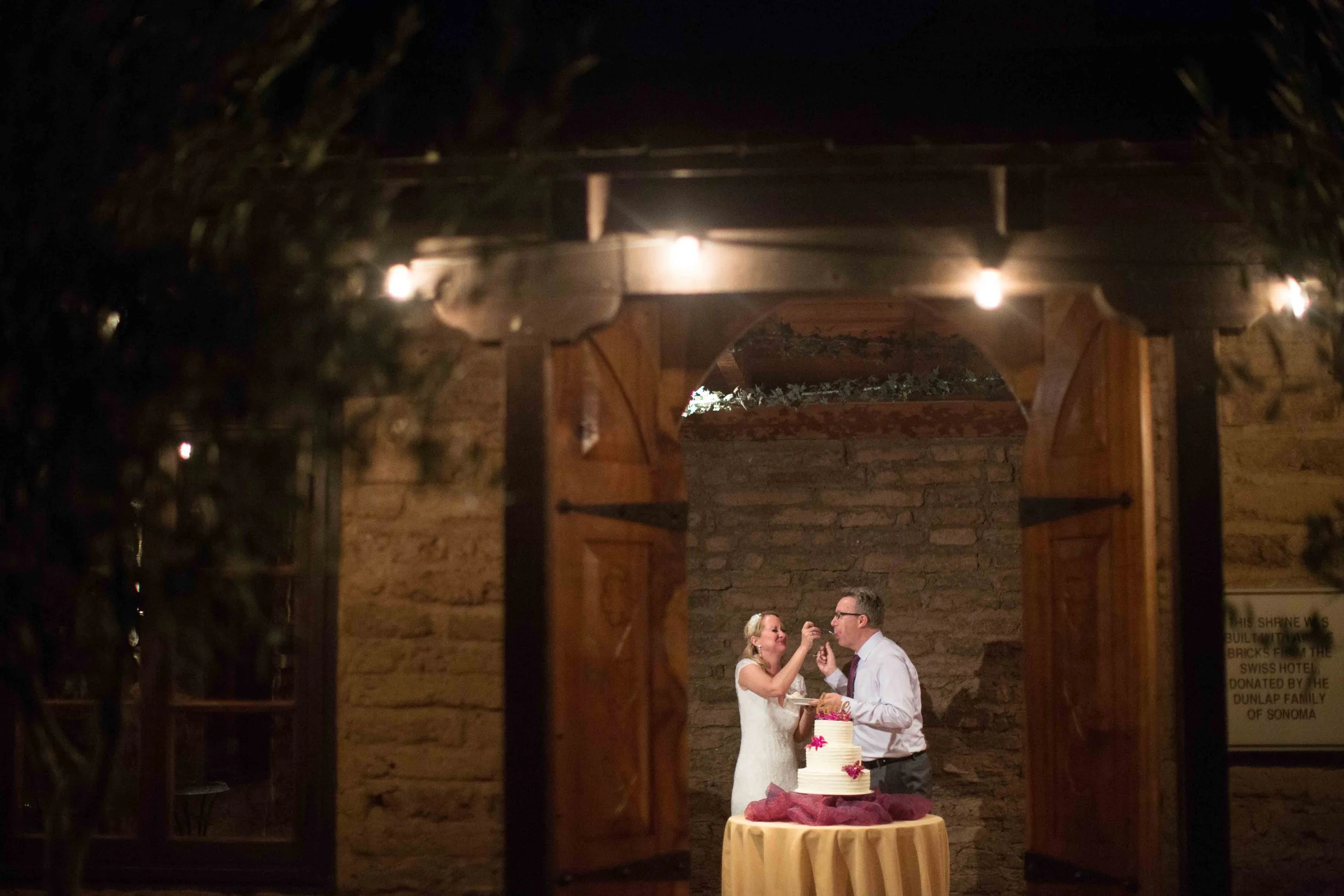 A bride and groom cutting a wedding cake at their reception in a rustic venue with a brick wall background, seen through a wooden-framed window with open shutters.
