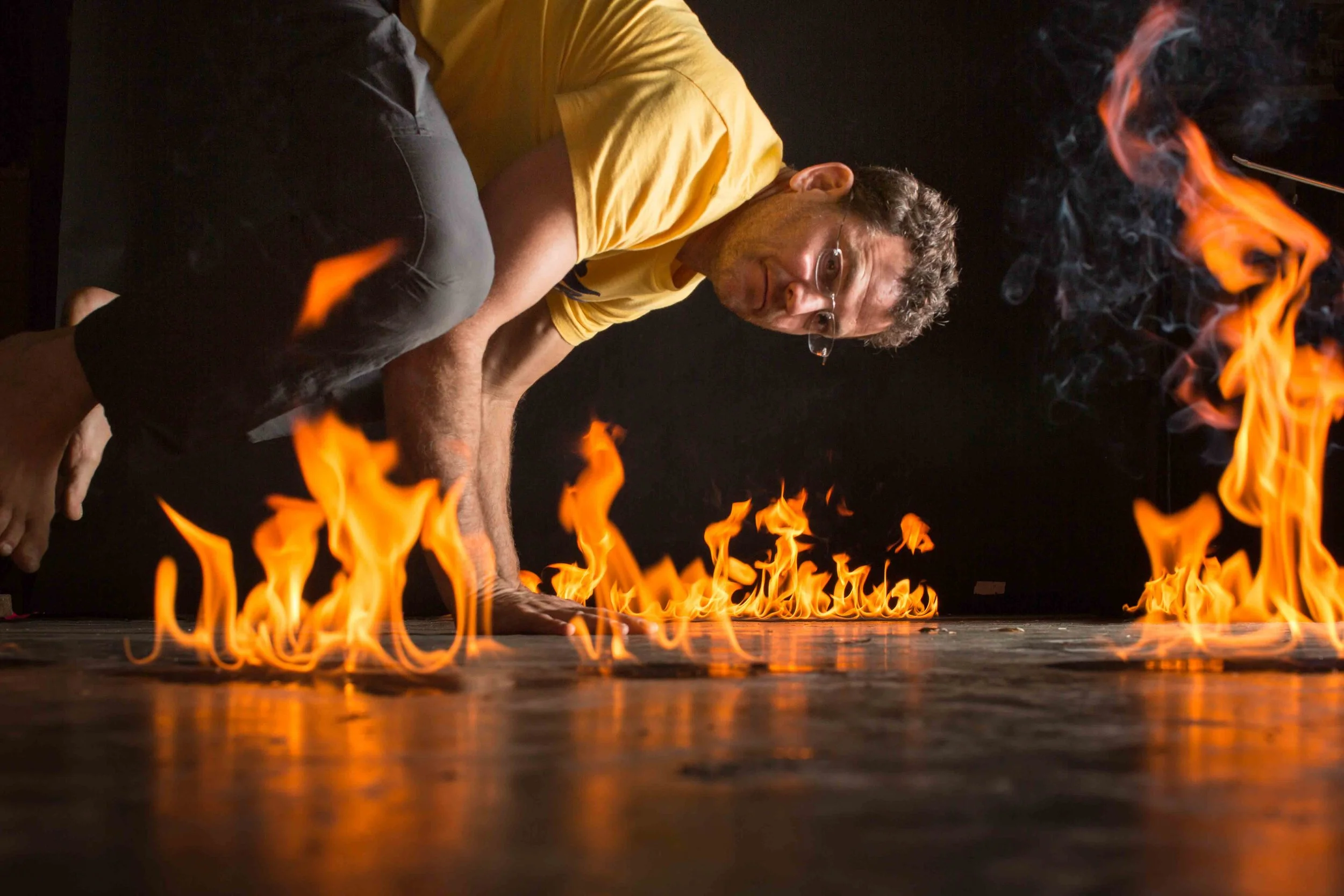 A man performing a dangerous stunt crawling on the floor amidst open flames in a dark room.