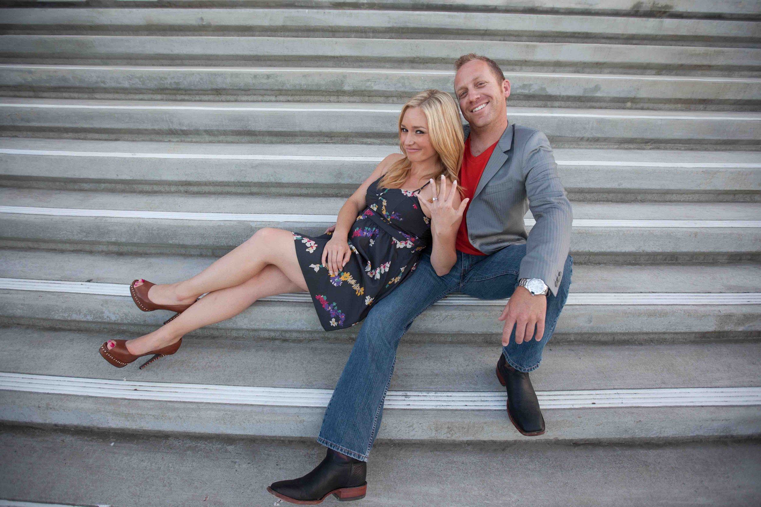 A man and woman sitting on outdoor concrete stairs, smiling and posing for the camera. The woman is wearing a sleeveless floral dress and high heels, while the man is dressed in a gray blazer, red shirt, jeans, and black boots.