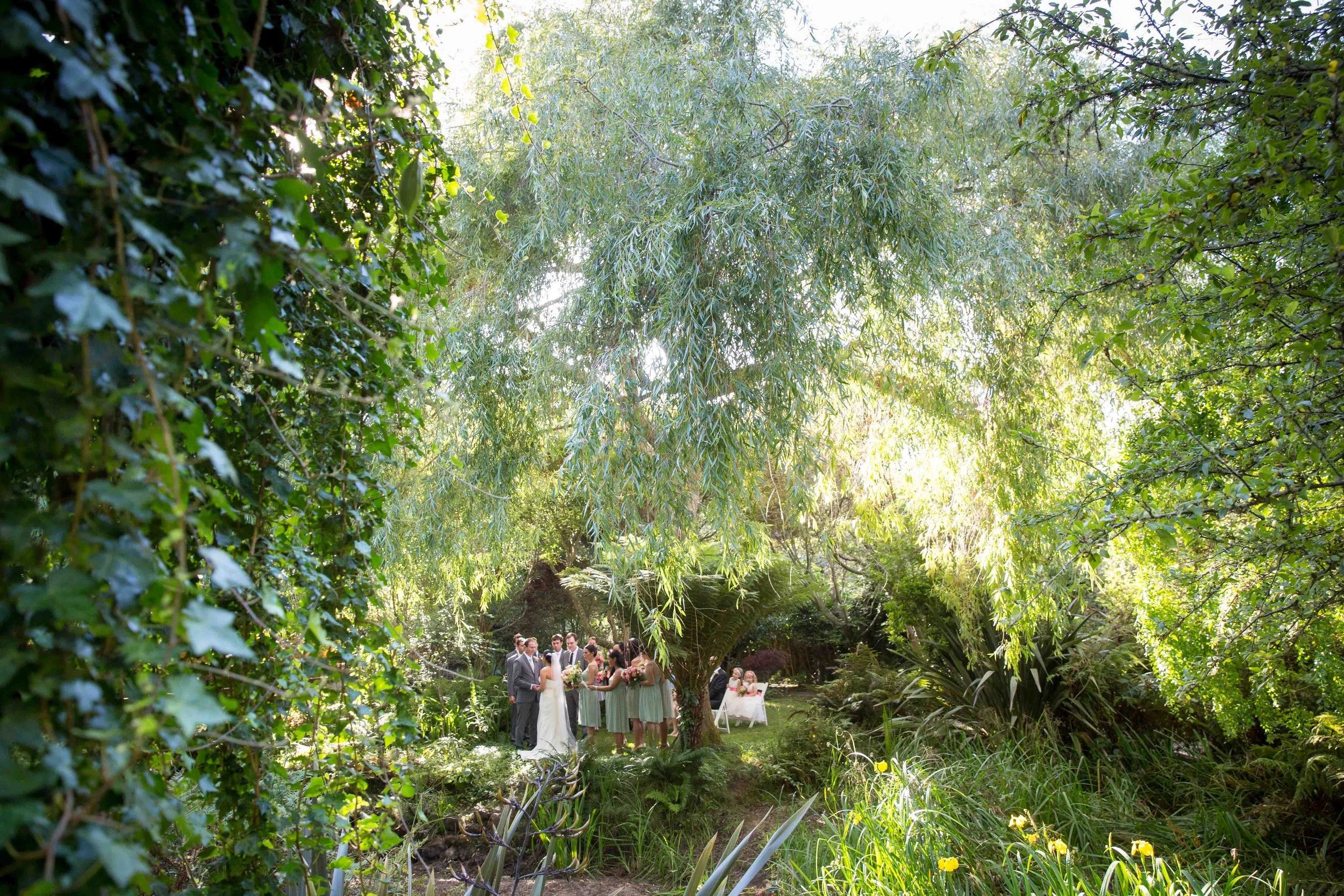 Wedding ceremony taking place outdoors in a lush green garden with large trees, plants, and sunlight filtering through the foliage. The bride and groom are standing together with their wedding party, some holding bouquets, in front of an arch or large tree at the center of the scene.