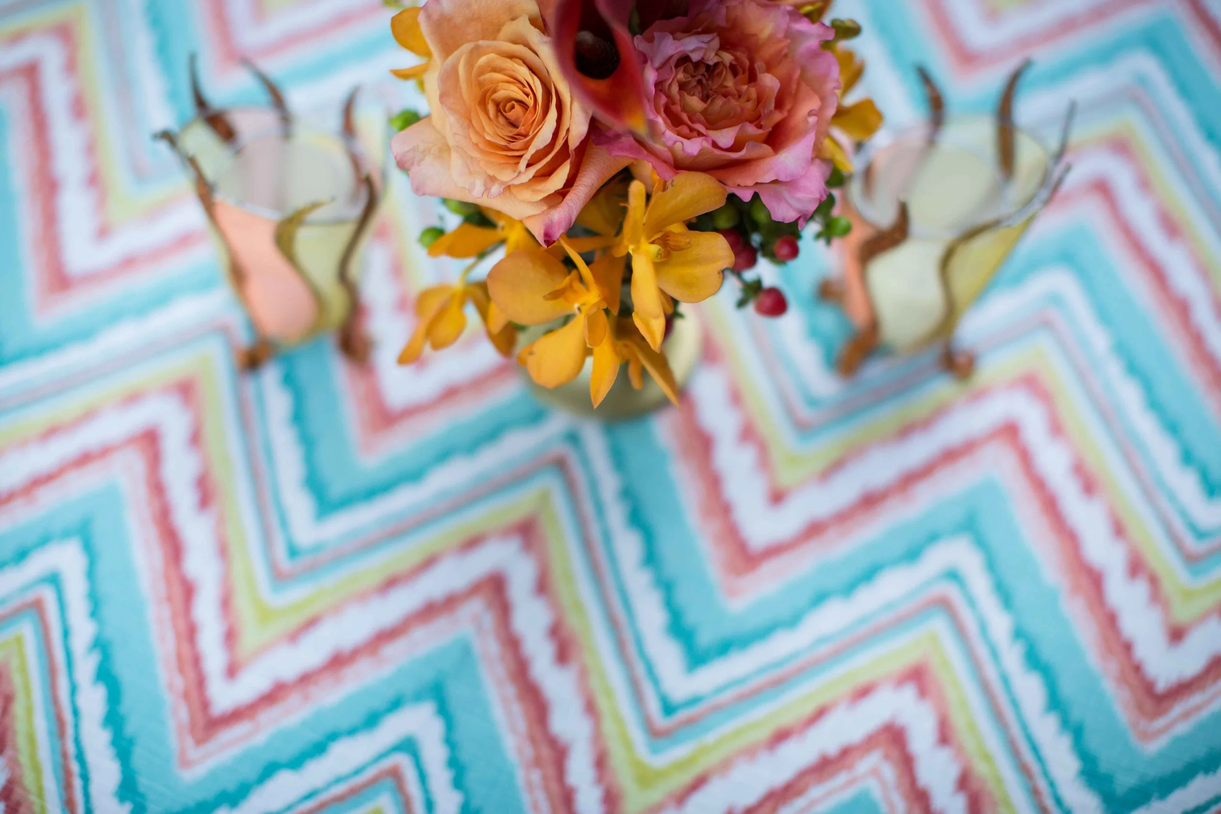 Top-down view of a floral arrangement with peach roses, yellow orchids, and pink flowers in a vase on a colorful zigzag patterned tablecloth.