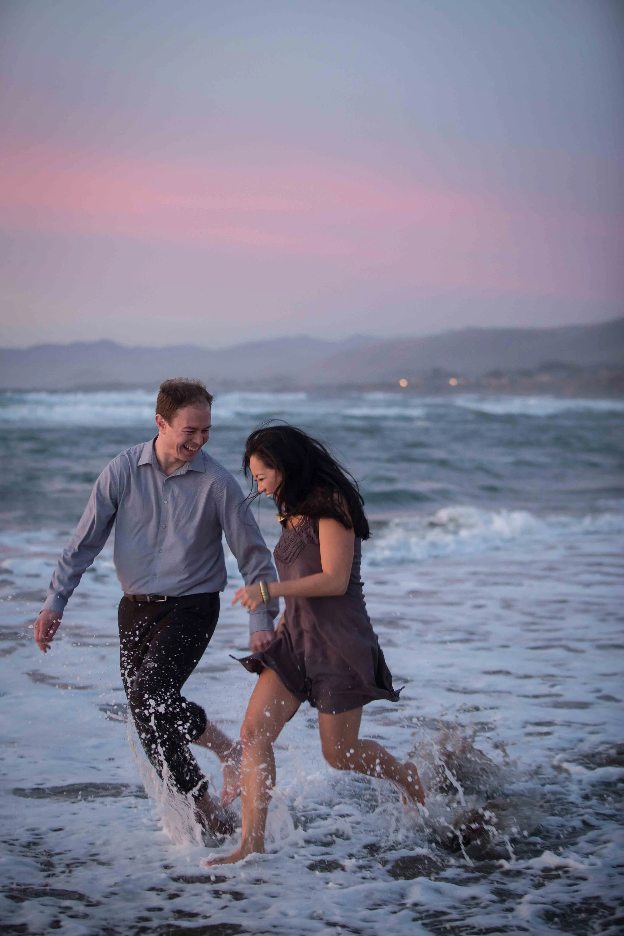 A man and a woman are playing and laughing in the ocean at sunset.