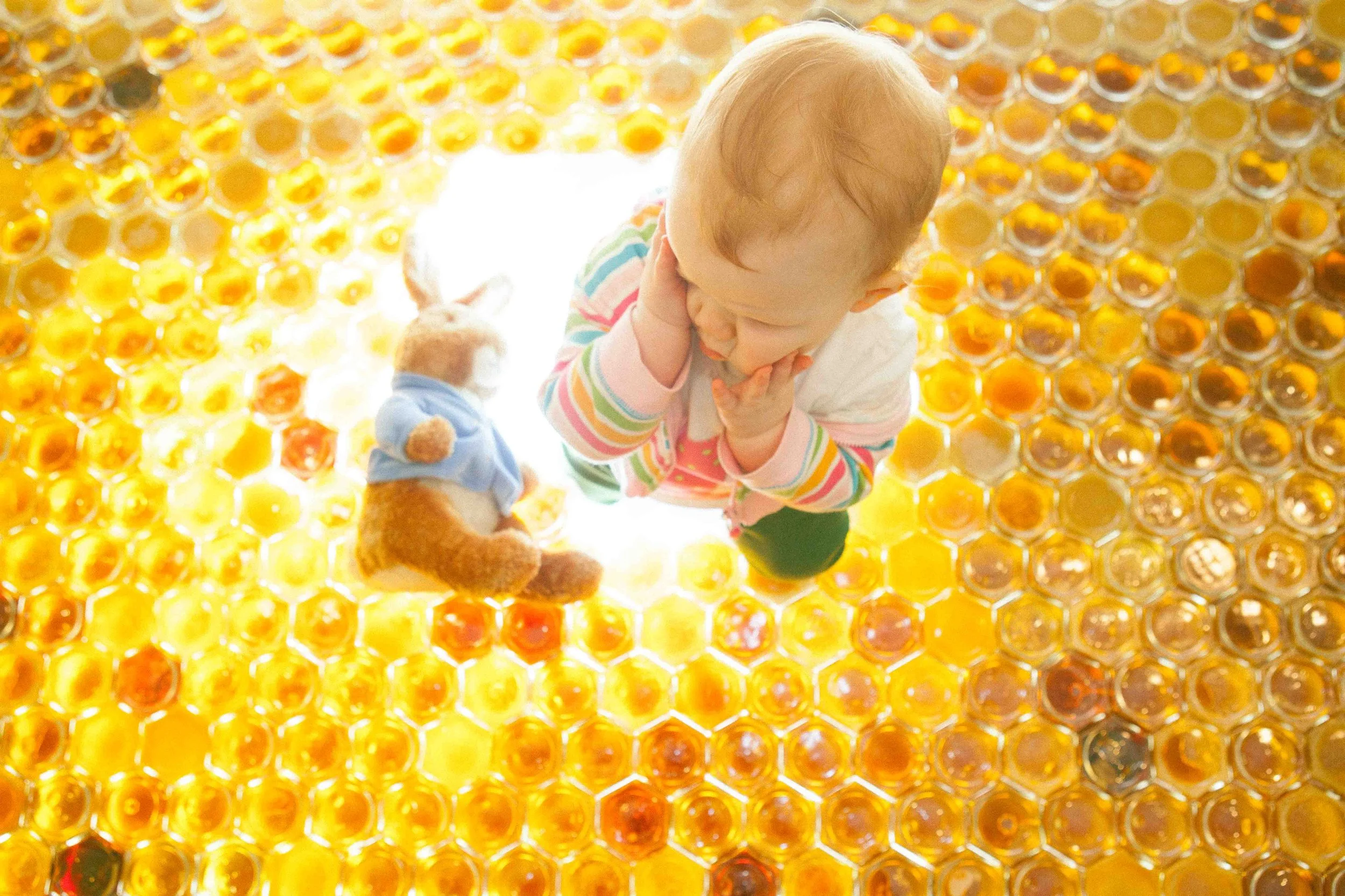 A young child with light hair and colorful striped shirt sitting on a honeycomb-patterned floor, looking down with one hand on their chin and the other on their cheek. A teddy bear with a blue outfit is laying on the floor nearby.