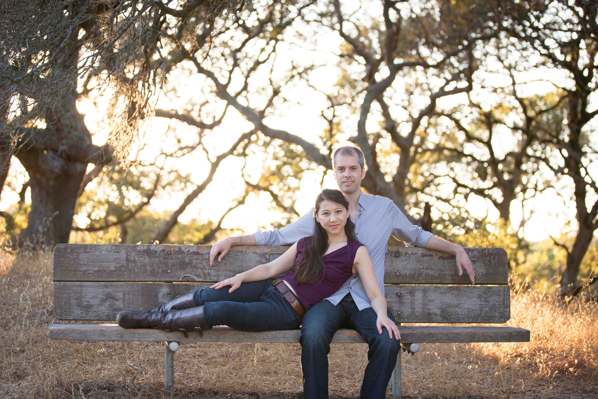 A couple sitting on a wooden park bench surrounded by autumn trees during sunset.