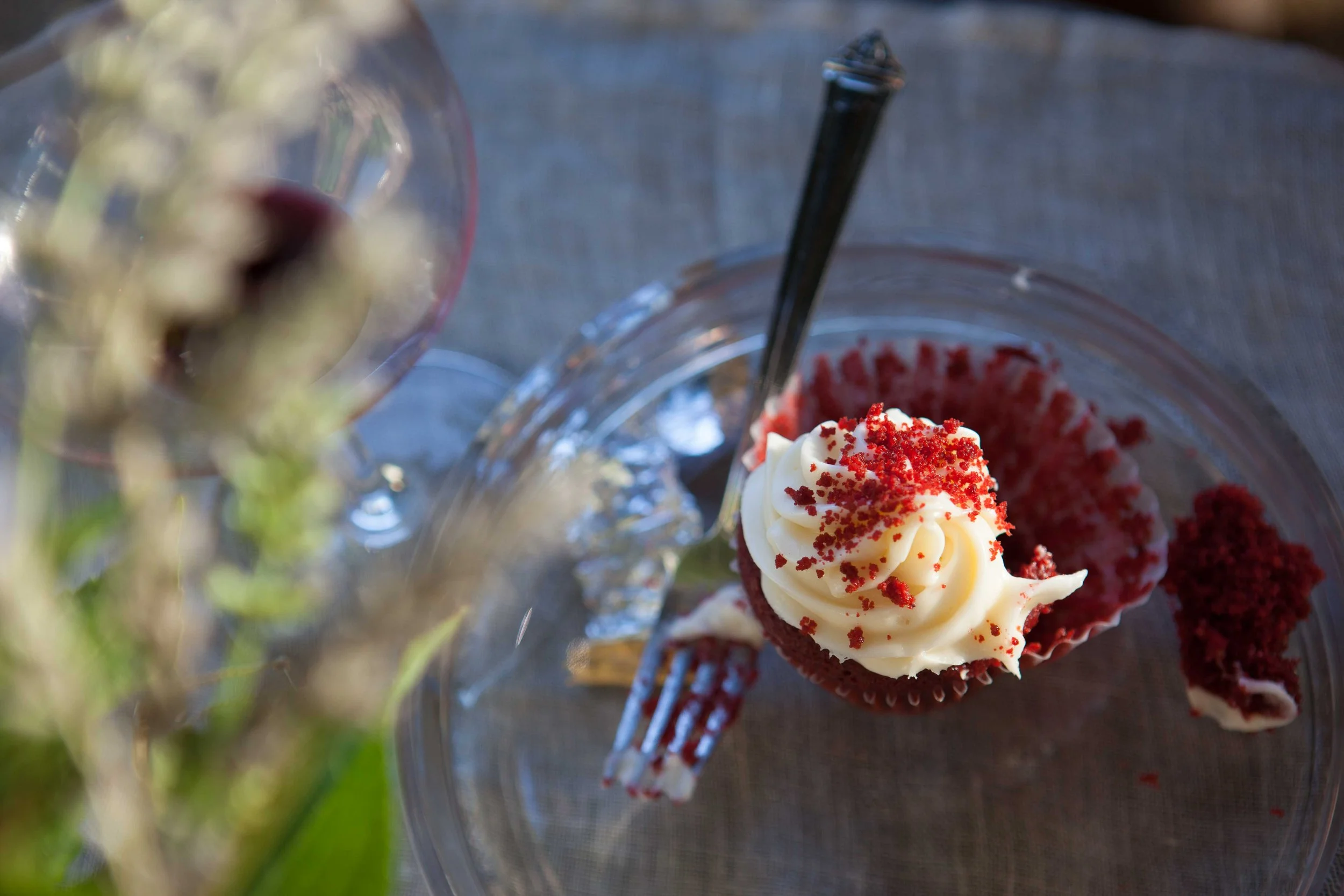 Red velvet cupcake with cream cheese frosting and red sprinkles on top, placed on a glass plate with a fork. In the foreground, a blurred glass with a dark beverage and flowers on a gray table.