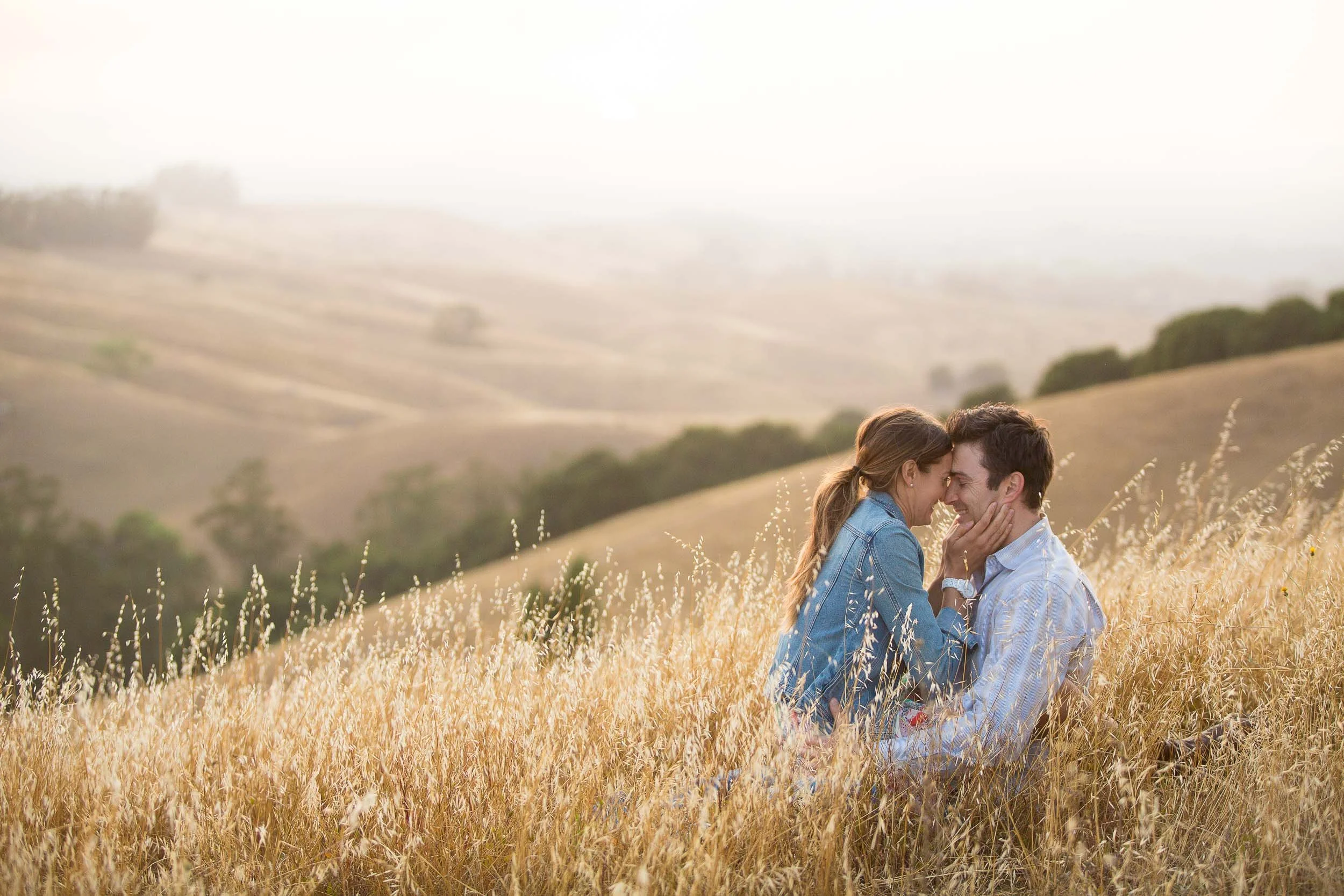 A couple sitting close together in a field of tall golden grass, touching foreheads and smiling, with rolling hills in the background on a hazy day.