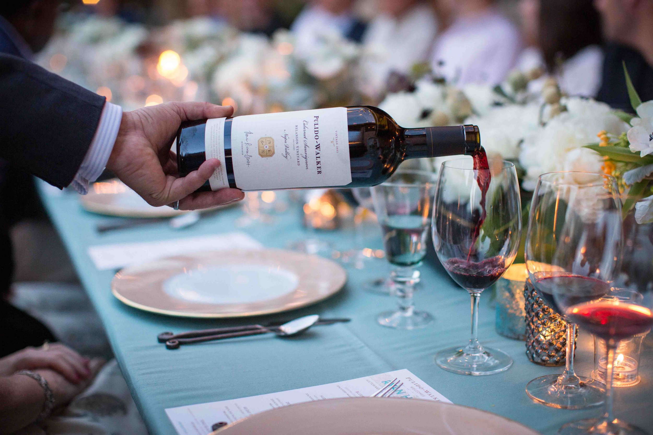 Person pouring red wine into a glass at a formal dinner table decorated with flowers and candles.