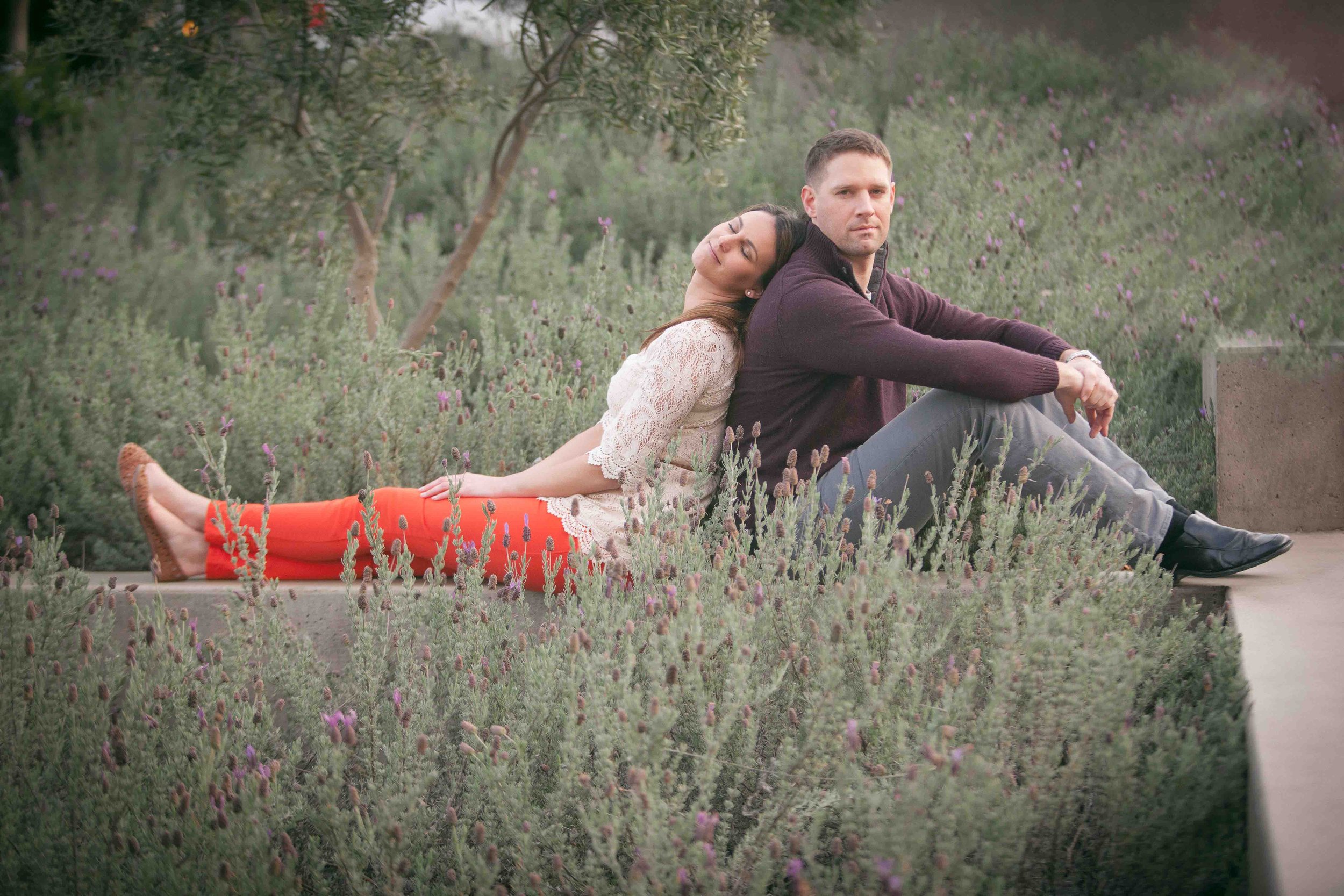 A young couple sits with their backs against each other on a concrete ledge in a garden with lavender and greenery; the woman is resting her head on the man's shoulder, both are looking in different directions.