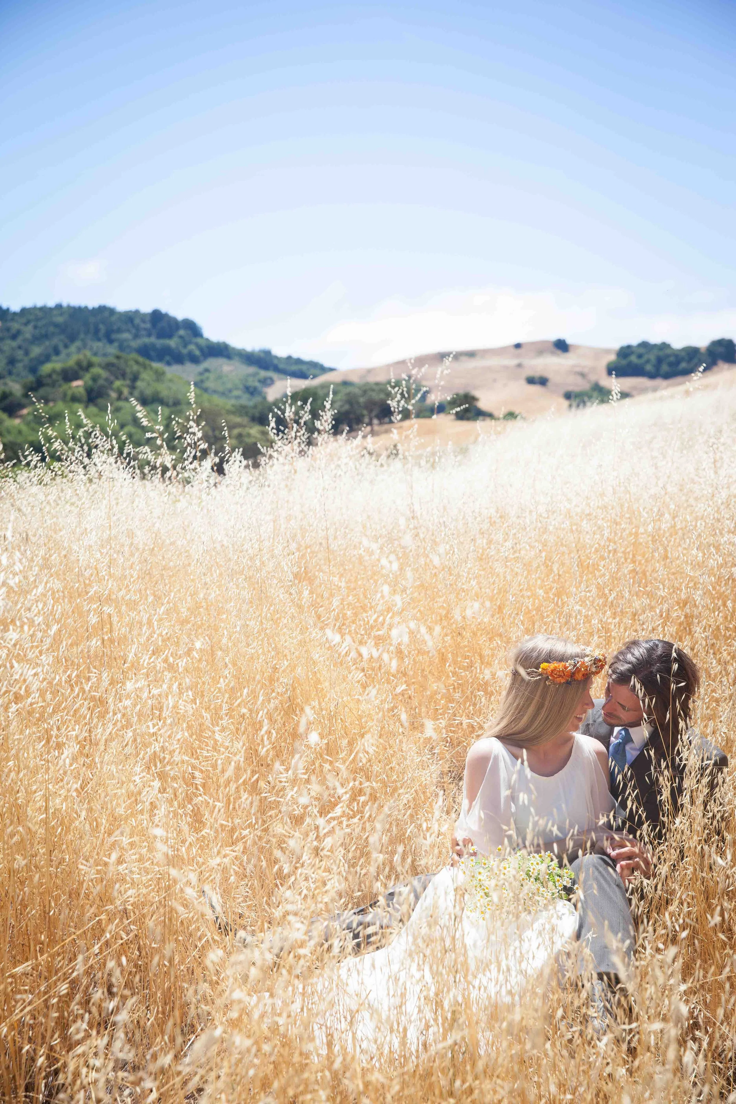 A couple sitting in a golden wheat field, with the woman wearing a white dress and floral crown, and the man in a suit, sharing an intimate moment under a bright blue sky with rolling hills in the background.