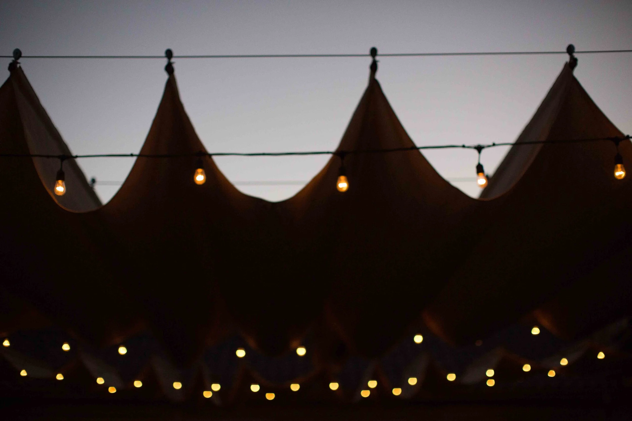 String of warm yellow string lights hanging under fabric canopies or tents at dusk or night.