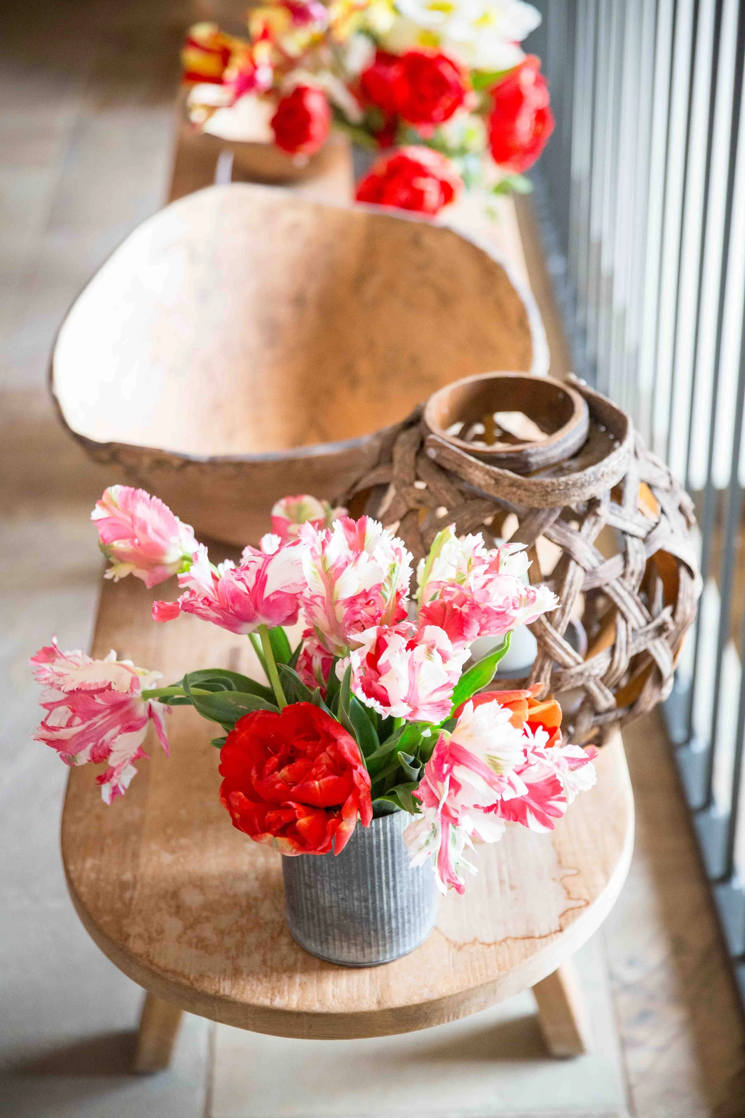 A small metal vase with pink and white tulips and a red tulip, placed on a wooden table with a decorative wicker lantern and a large wooden bowl in the background.