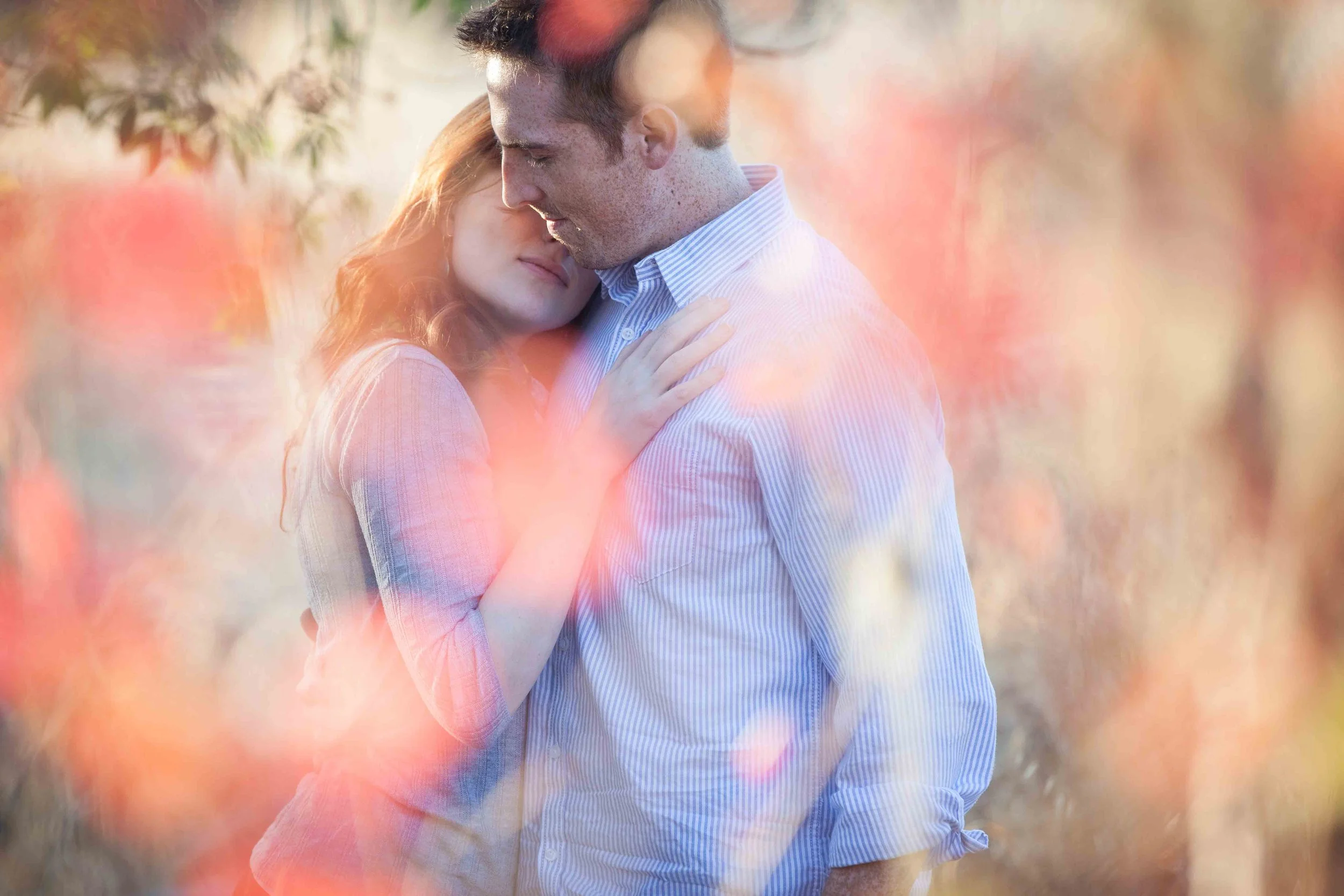 A couple embracing softly outdoors during sunset, surrounded by pink and orange blurred foliage.