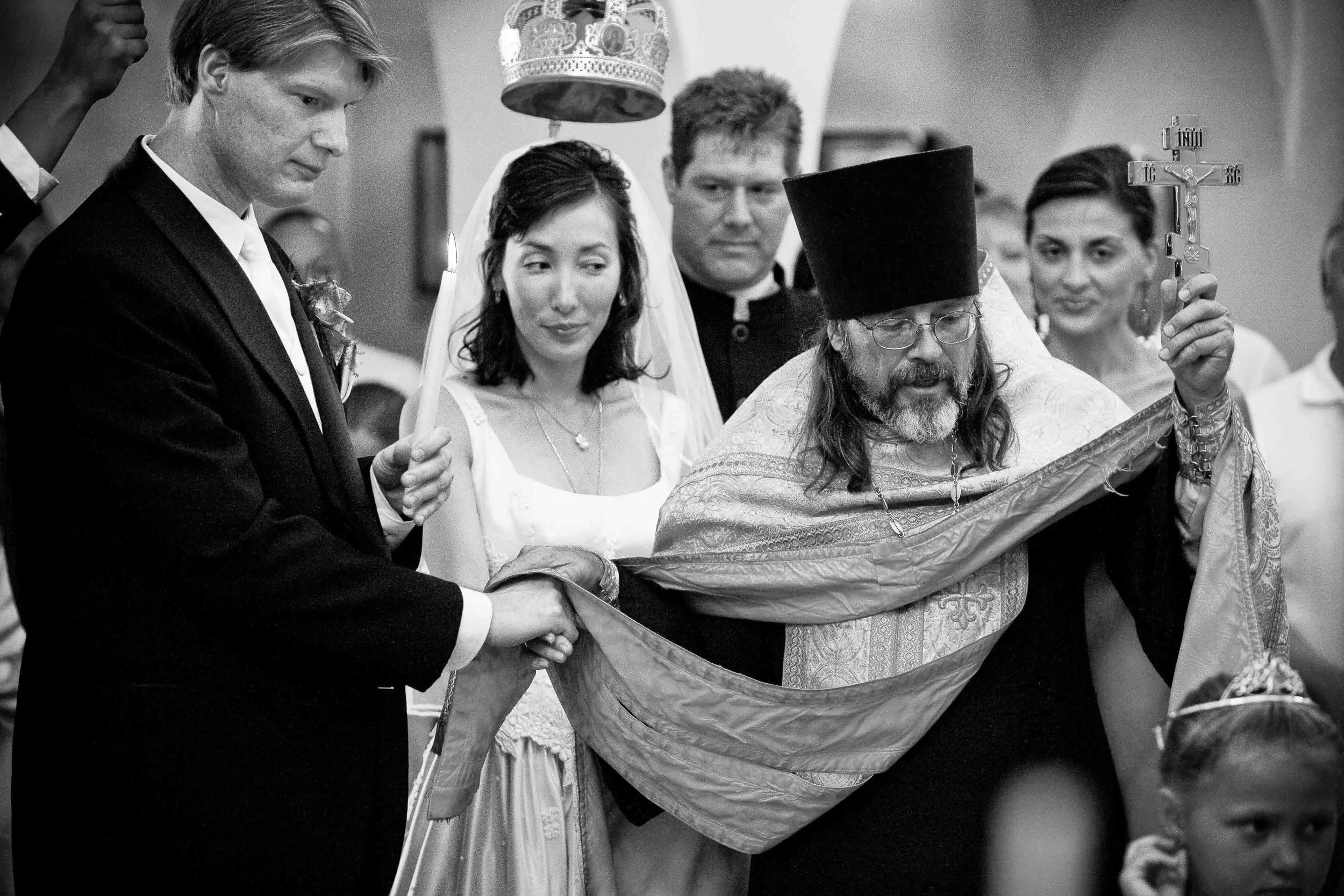 A black-and-white photo of a wedding ceremony inside a church with a priest turning a couple around with a cloth, surrounded by an officiant, the bride and groom, and other guests.