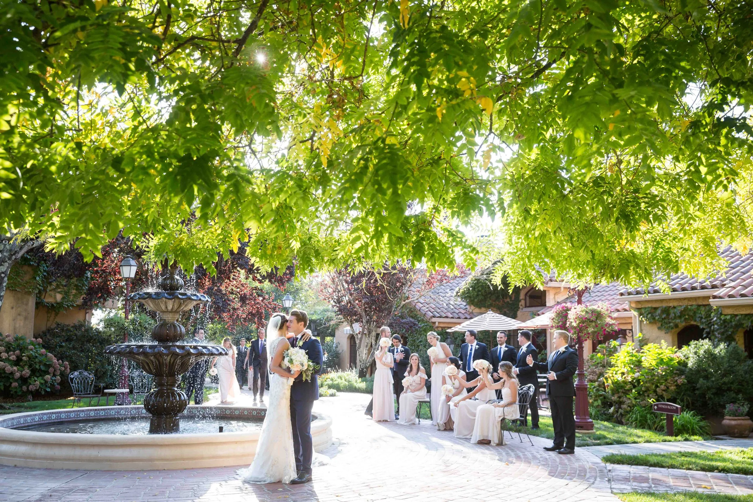 A wedding ceremony taking place outdoors in a garden with a fountain, surrounded by trees and flowering bushes. The bride and groom are standing under a large tree, with bridesmaids seated nearby holding bouquets, and groomsmen standing in a line.