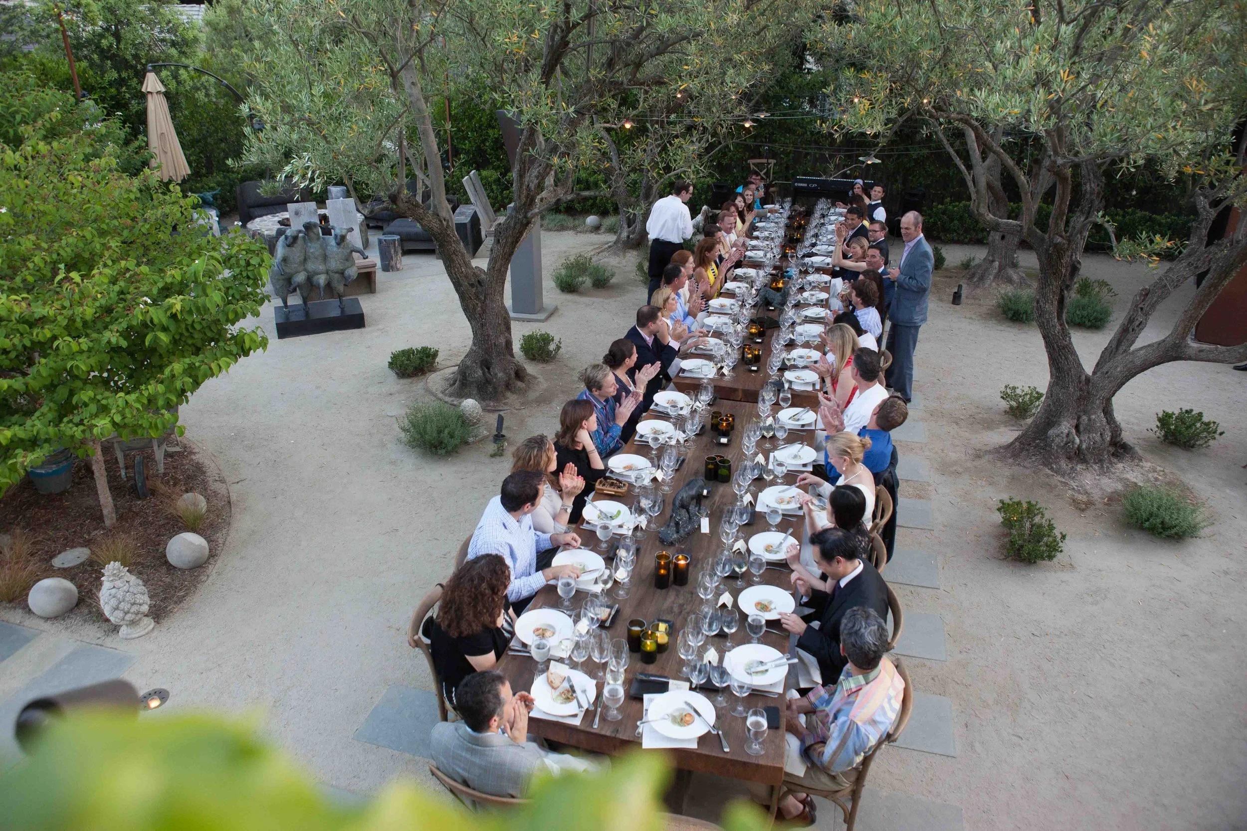 A large group of people gathered around a long rectangular wooden dining table outdoors, set with plates, glasses, and candles, under a canopy of trees during evening.