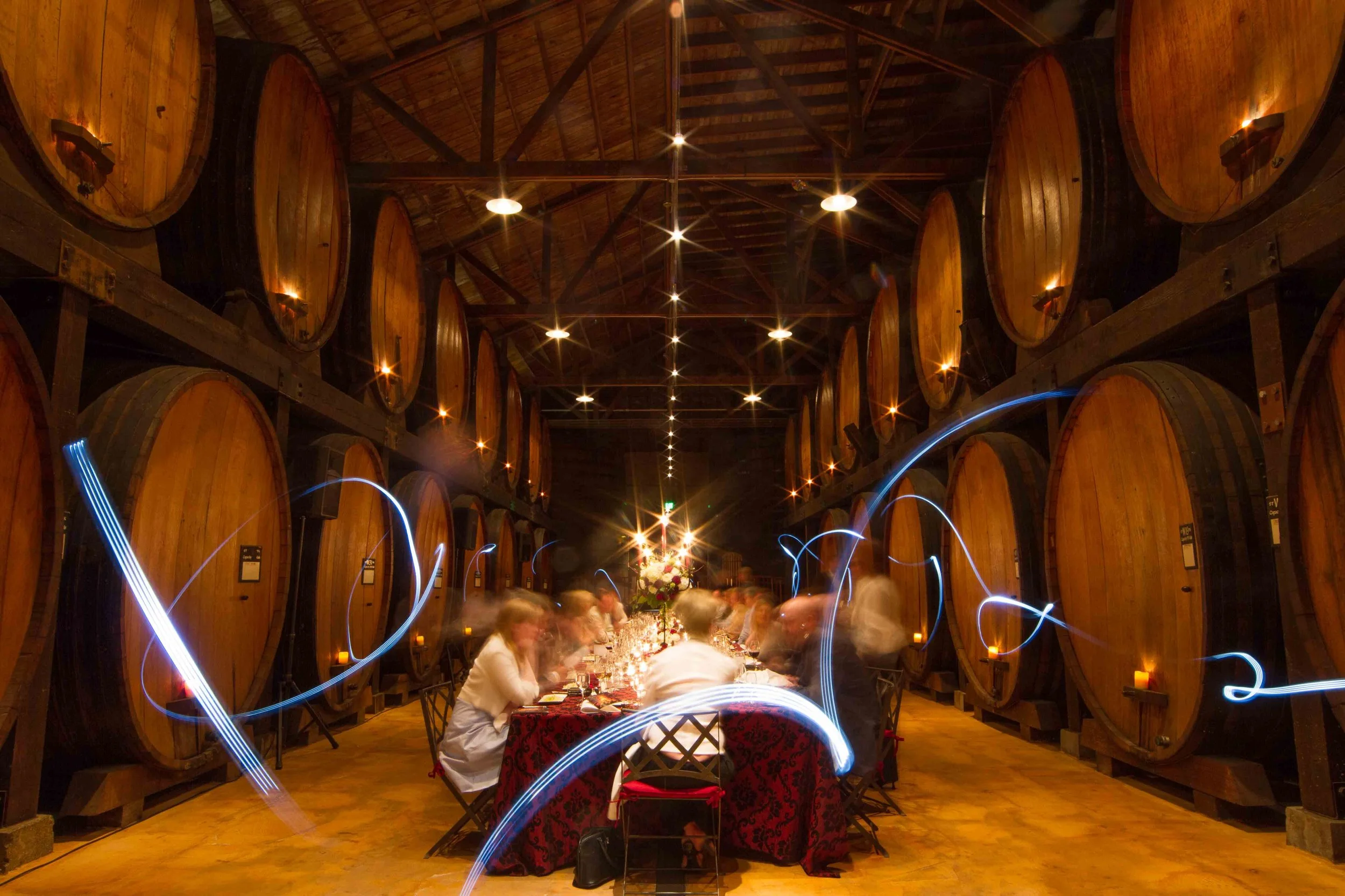 An indoor wine cellar or tasting room with large wooden wine barrels lining the walls, a long table with people dining, and light streaks from long exposure photography.
