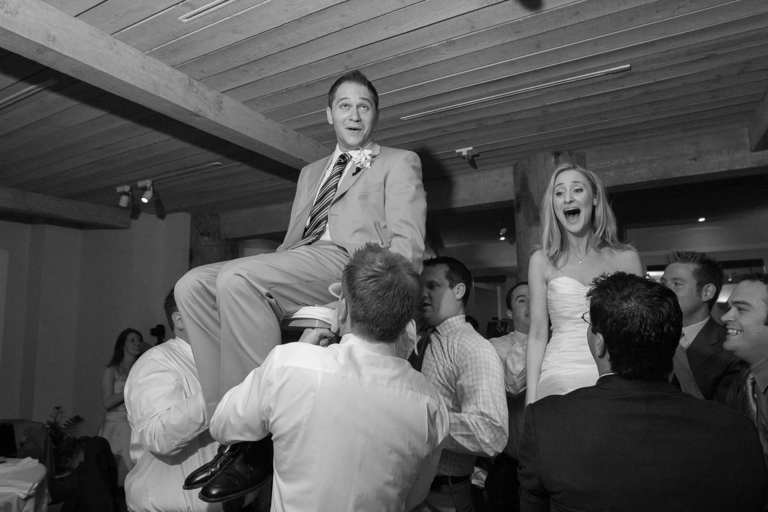 A man in a suit and a woman in a wedding dress sit on chairs in the air, being lifted by a group of people at a wedding reception.