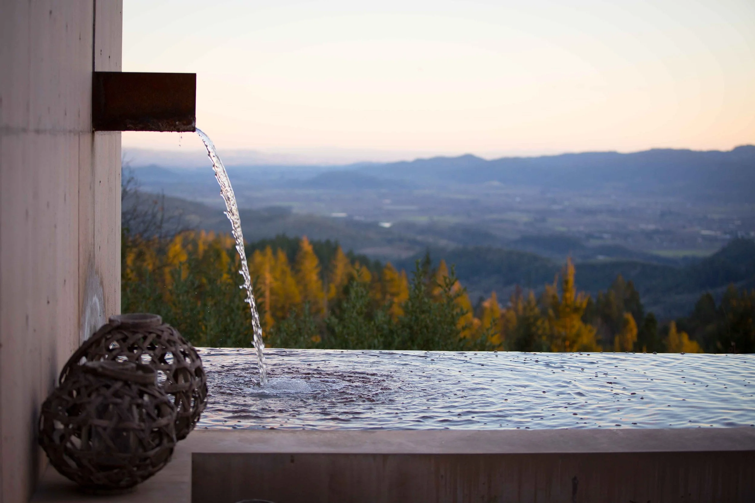 A wooden outdoor water feature with a small waterfall, overlooking a landscape of rolling hills and trees with autumn foliage at sunset.
